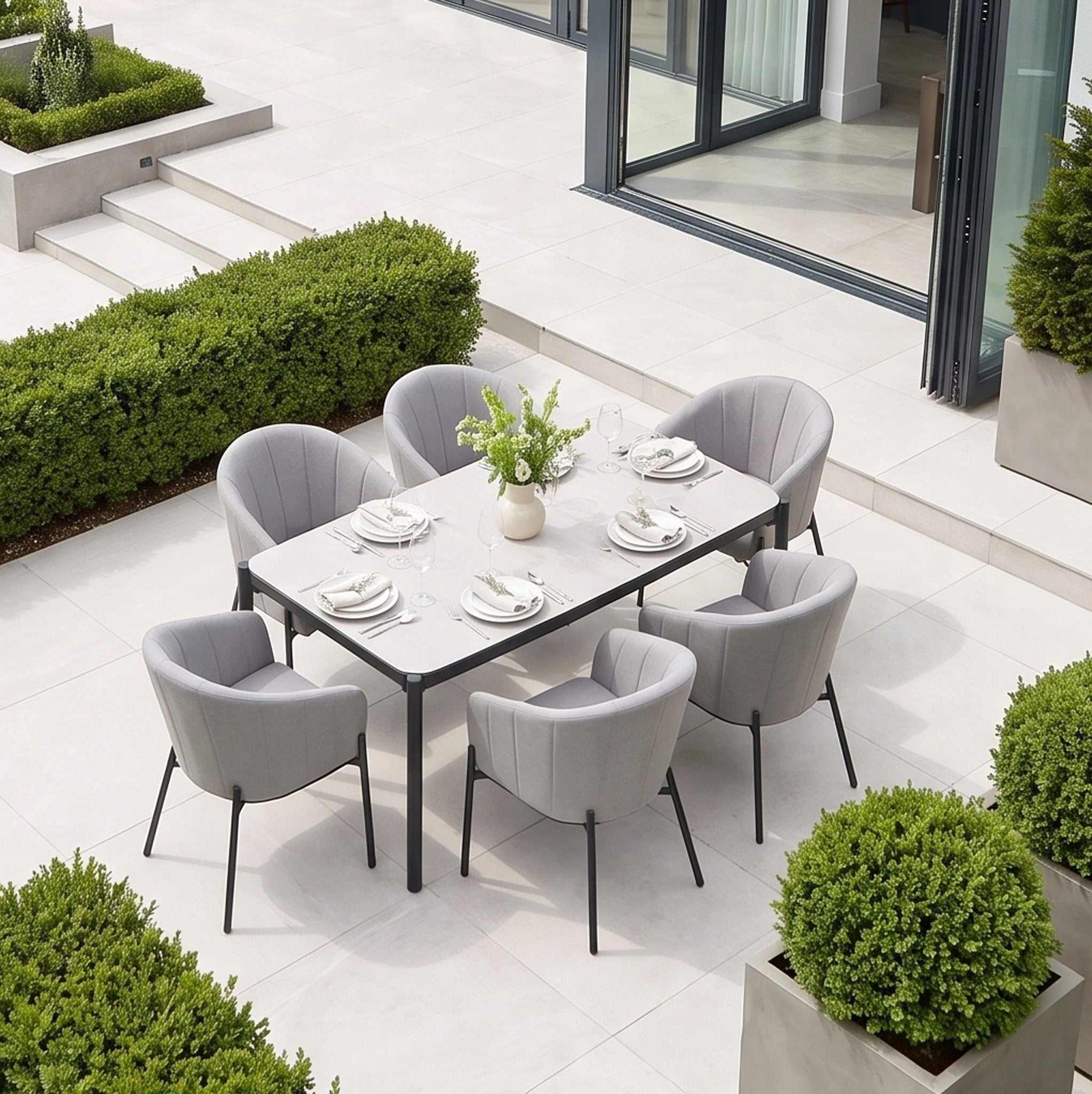 Elevate your garden dining experience with the Kai 6 Seat Rectangular Dining Set in Light Grey, featuring a powder-coated aluminium frame, six elegant gray chairs, white tableware, and vibrant green plants on a stylish tiled patio.