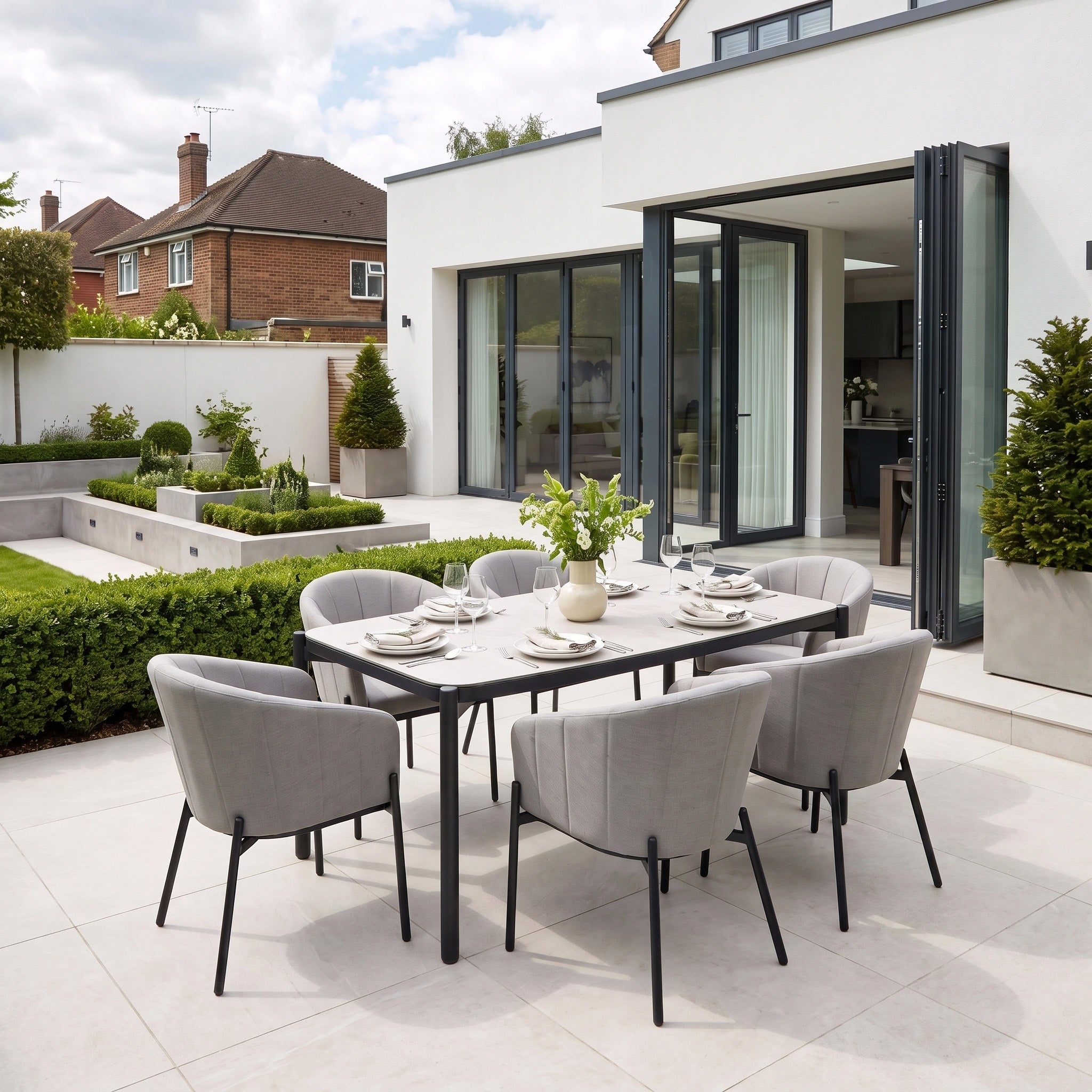 Modern outdoor dining area featuring the Kai 6 Seat Rectangular Dining Set in Light Grey, with gray chairs and a set table, situated in a landscaped backyard beside a white house. This stylish garden dining set is crafted with a durable powder-coated aluminium frame, making it perfect for al fresco gatherings.