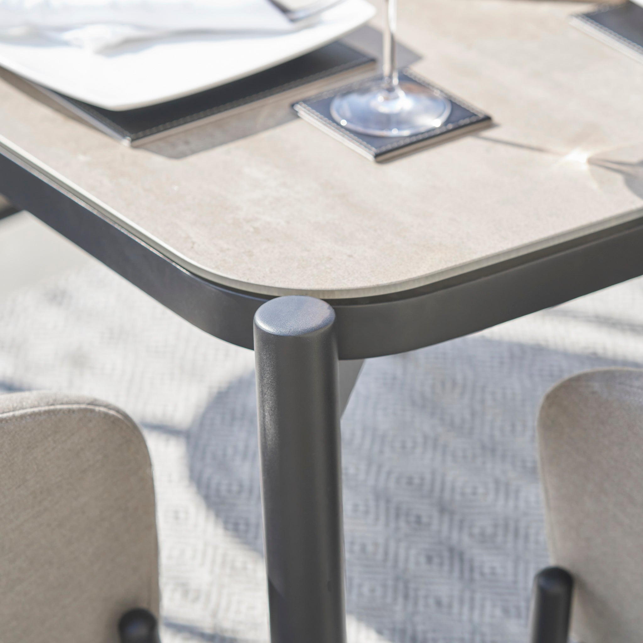 Close-up of the Kai 6 Seat Rectangular Dining Set in Light Grey, featuring a modern table corner with a glass, placemat, and two beige chairs on a patterned rug—perfect for complementing your outdoor dining set or enhancing your garden furniture collection.
