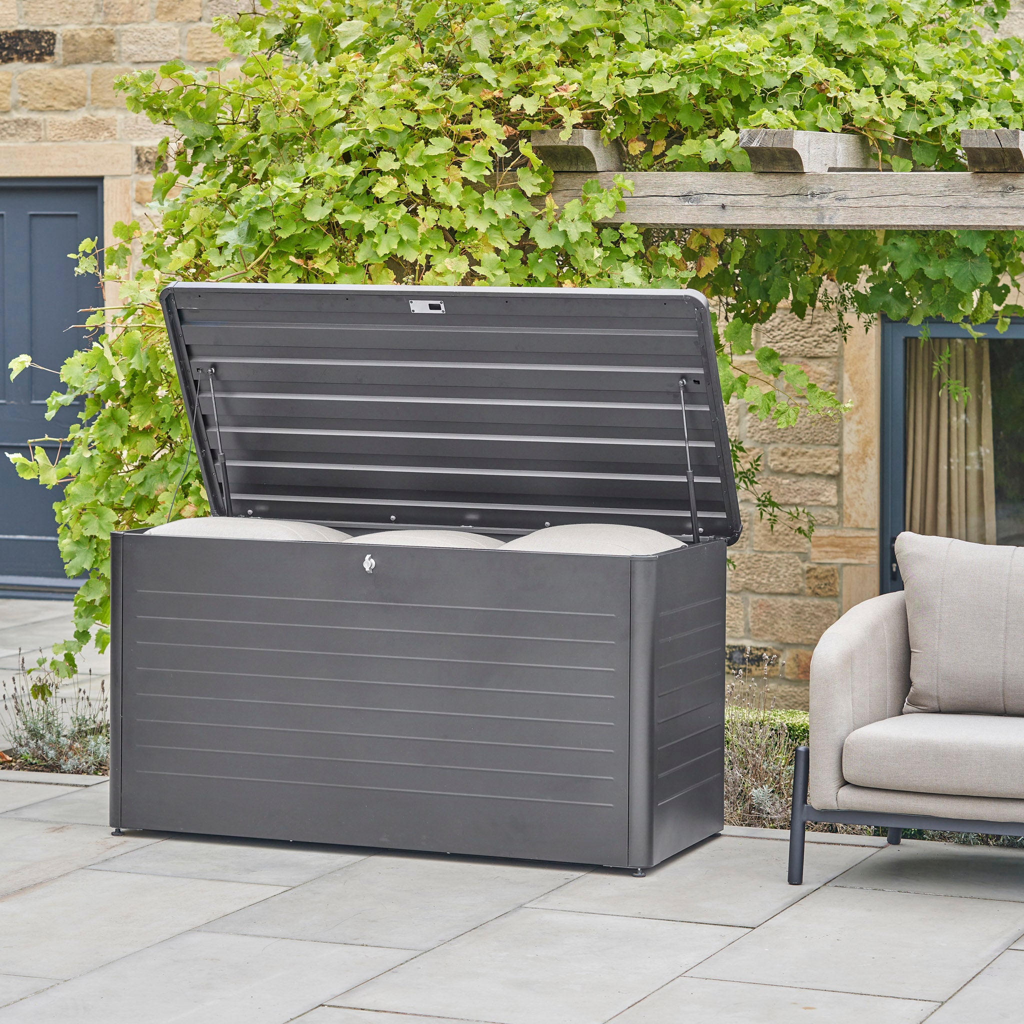 Pacific Lifestyle - Rhode Island Large Outdoor Cushion Box in Anthracite: a galvanized steel storage box with open lid neatly stores cushions beside a gray patio chair and green plants.