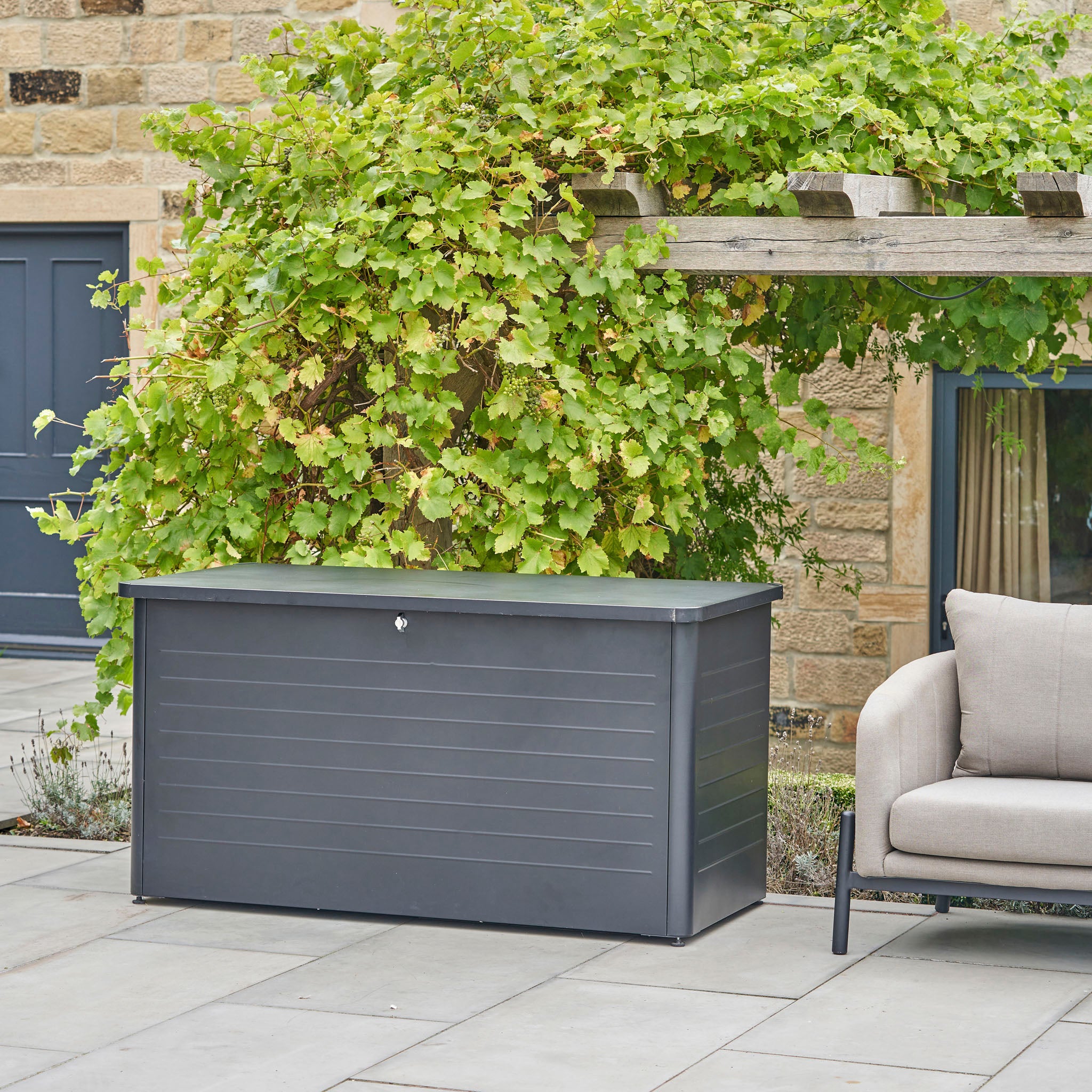A Pacific Lifestyle - Rhode Island Large Outdoor Cushion Box in Anthracite sits on a patio beside a sofa and lush vine-covered pergola, providing stylish outdoor storage for any space.