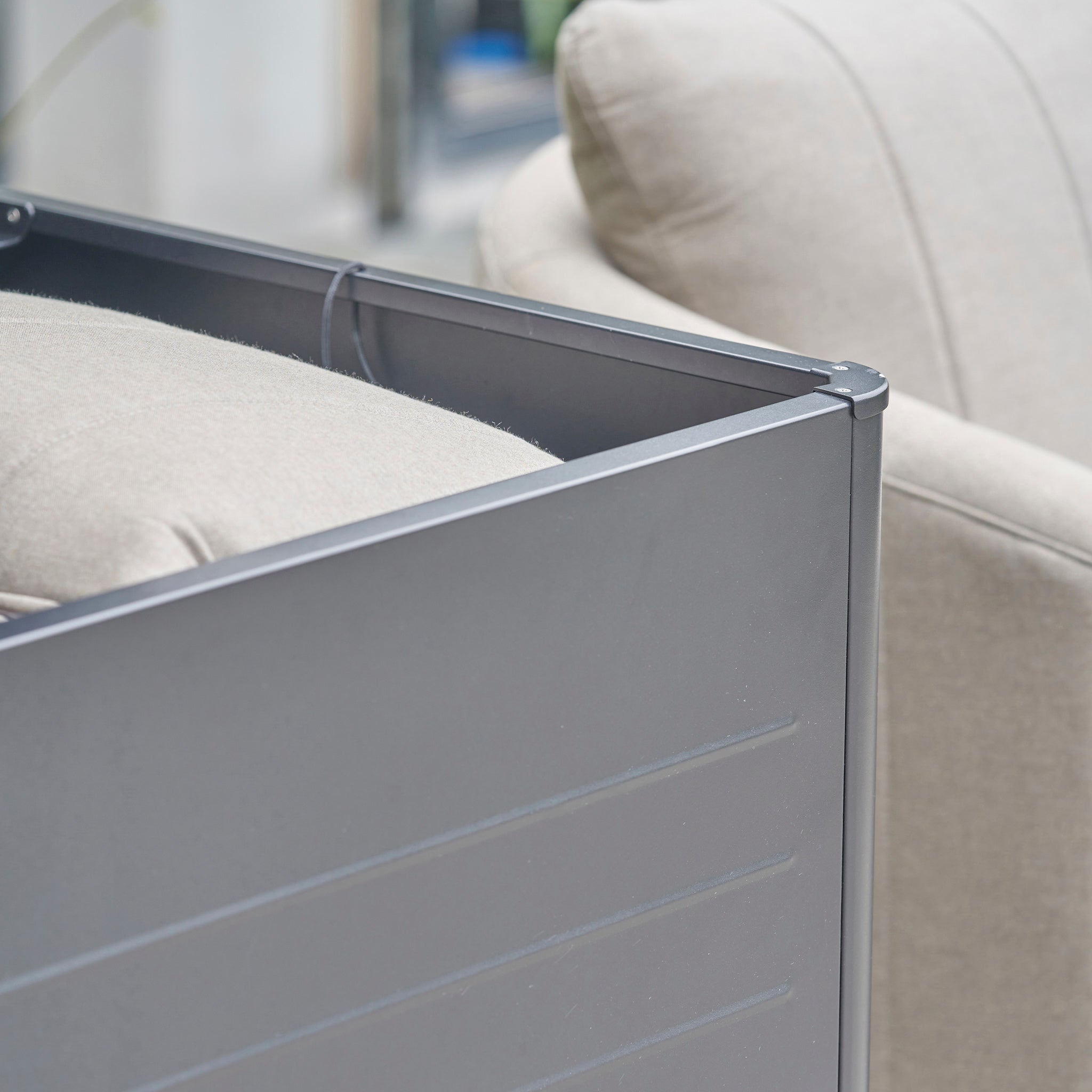 Close-up of a beige cushioned chair beside a gray galvanized steel panel, similar to the Pacific Lifestyle - Rhode Island Large Outdoor Cushion Box in Anthracite, shown indoors.