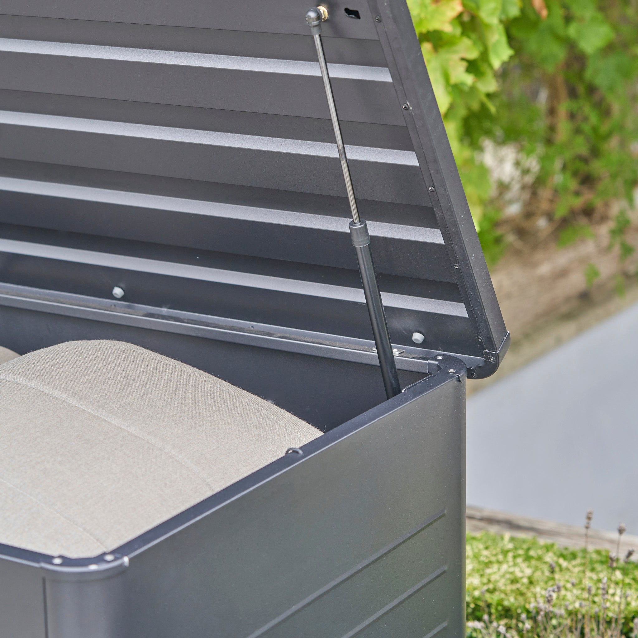 The Pacific Lifestyle - Rhode Island Large Outdoor Cushion Box in Anthracite features a black galvanised steel design with a beige cushion inside and its lid propped open.