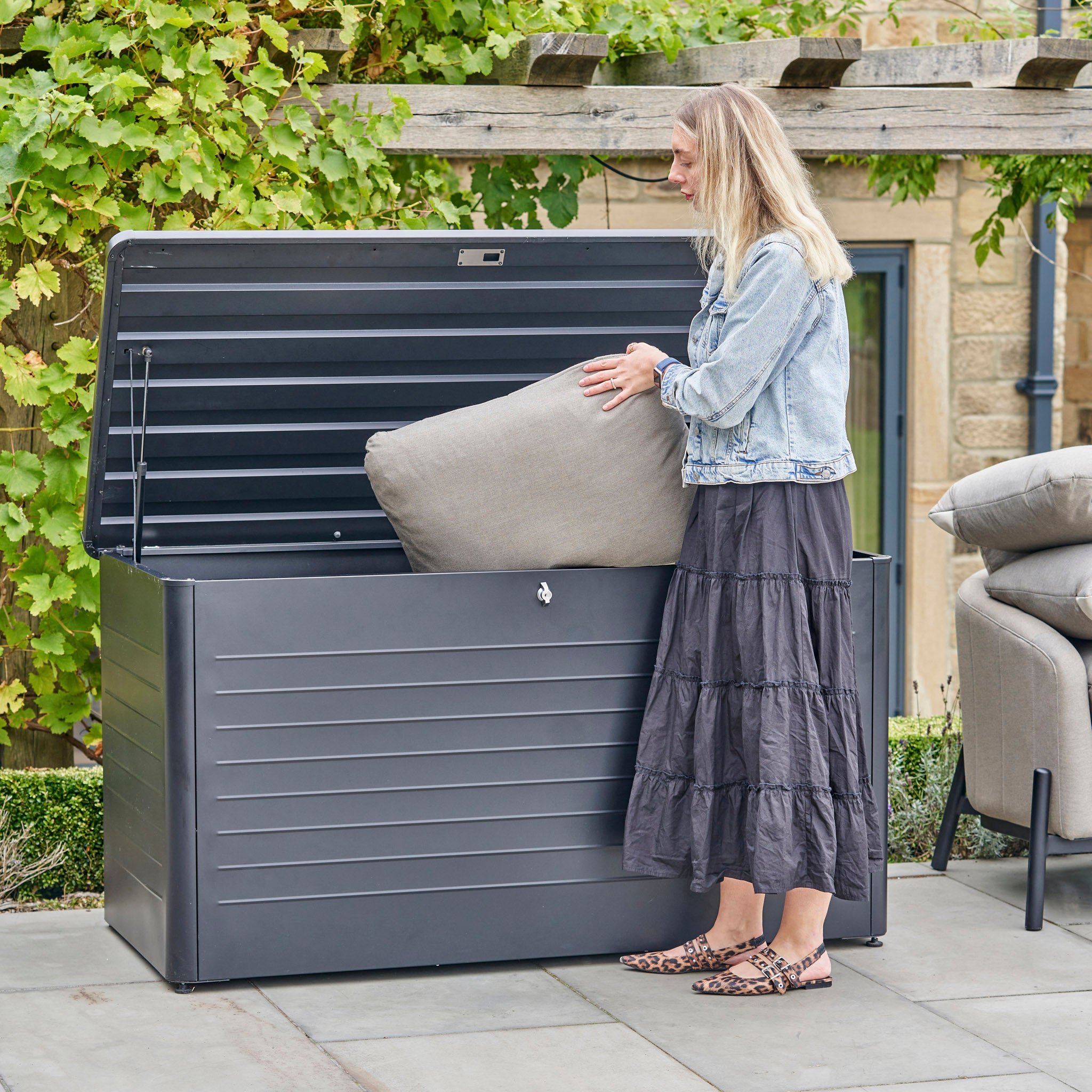 A woman places a large cushion into the Pacific Lifestyle - Rhode Island Large Outdoor Cushion Box in Anthracite on a patio near a vine-covered pergola, keeping her patio essentials protected and organized.