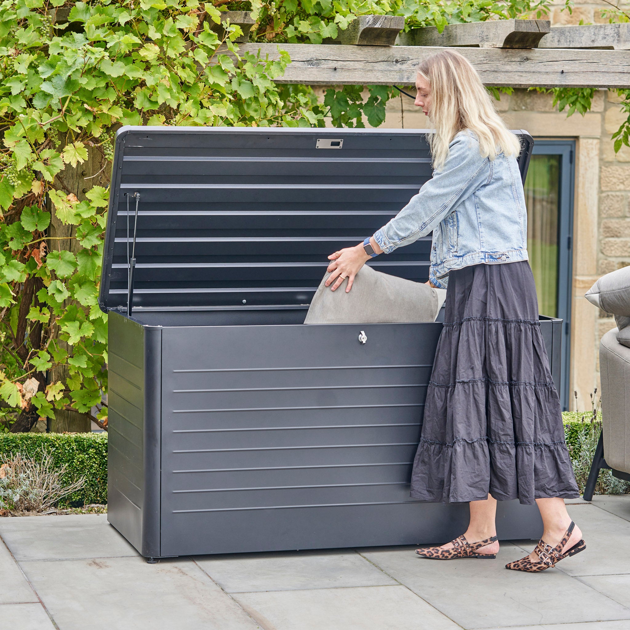 A woman places a cushion into the Pacific Lifestyle - Rhode Island Large Outdoor Cushion Box in Anthracite on the patio, surrounded by greenery. This spacious box keeps your patio essentials protected and organized all year round.
