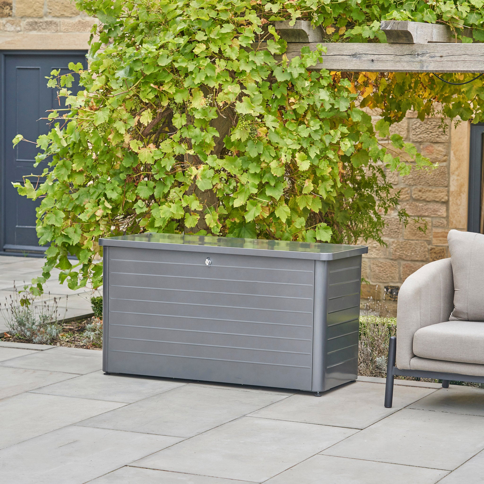 The Pacific Lifestyle Rhode Island Small Outdoor Cushion Box in Anthracite sits on a stone patio beside a sofa, with leafy green vines in the background.