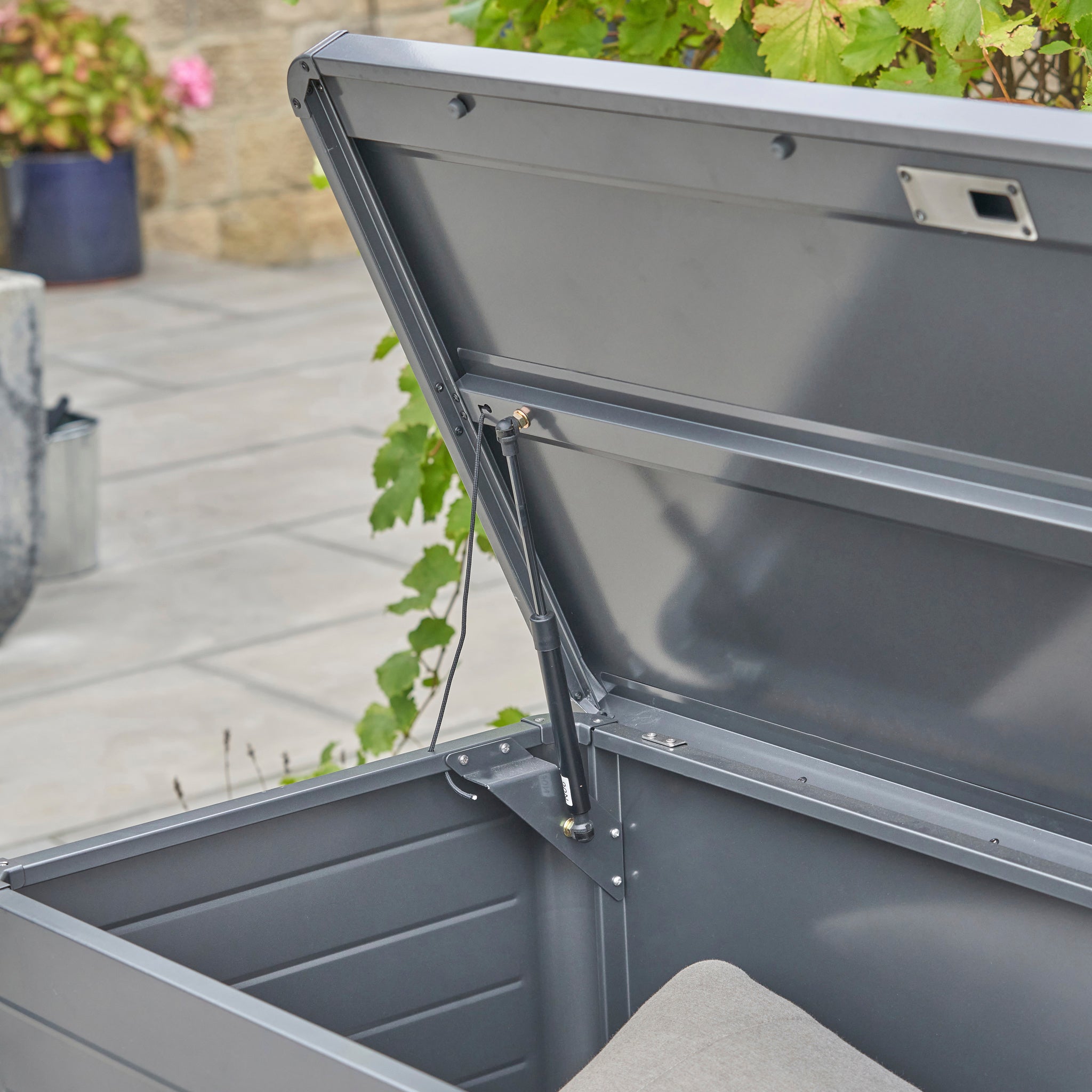 The Pacific Lifestyle - Rhode Island Small Outdoor Cushion Box in Anthracite, made of galvanised steel, sits on a patio amid lush plants with a partially visible cushion inside.