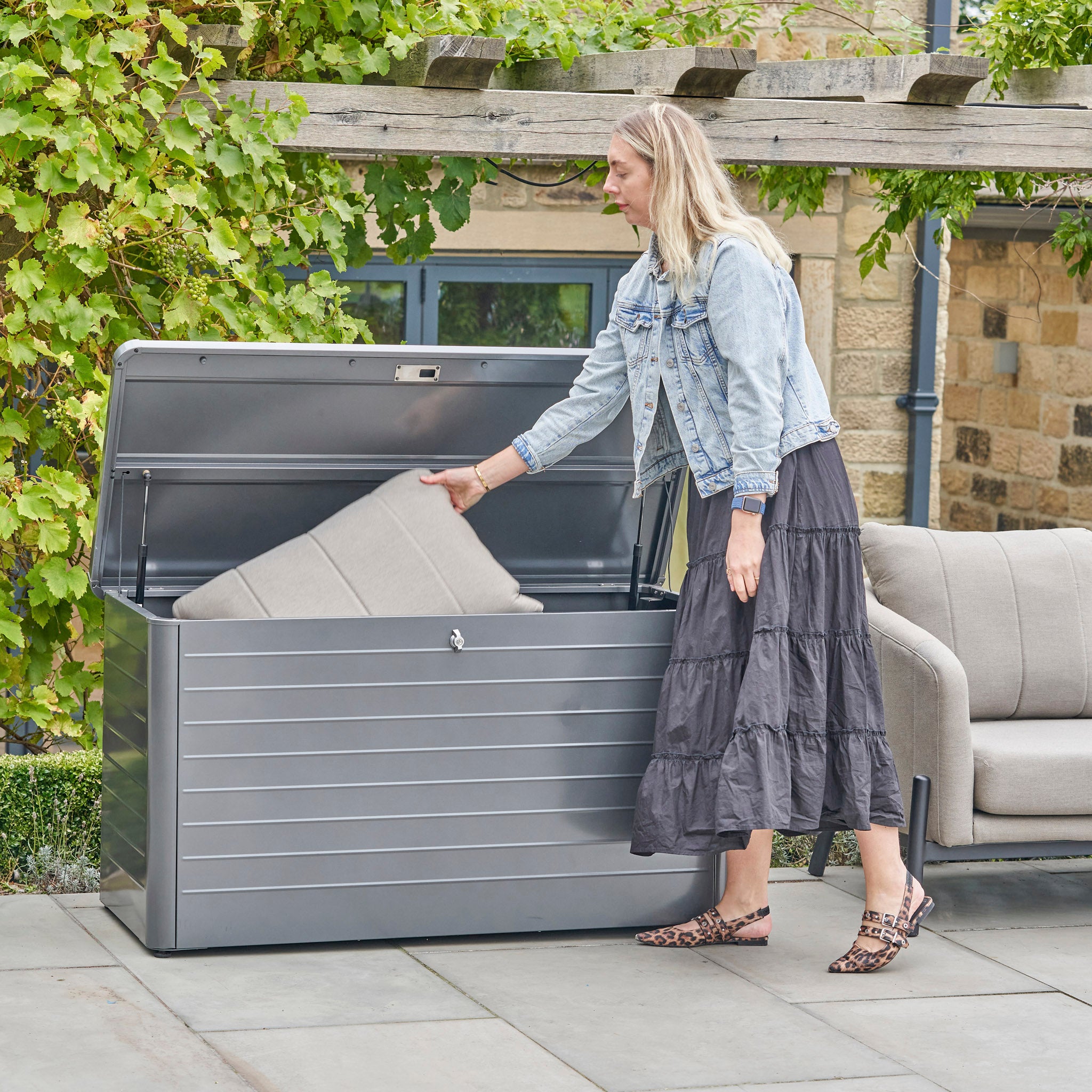 A woman places a cushion into the Pacific Lifestyle Rhode Island Small Outdoor Cushion Box in Anthracite on a patio next to a sofa.