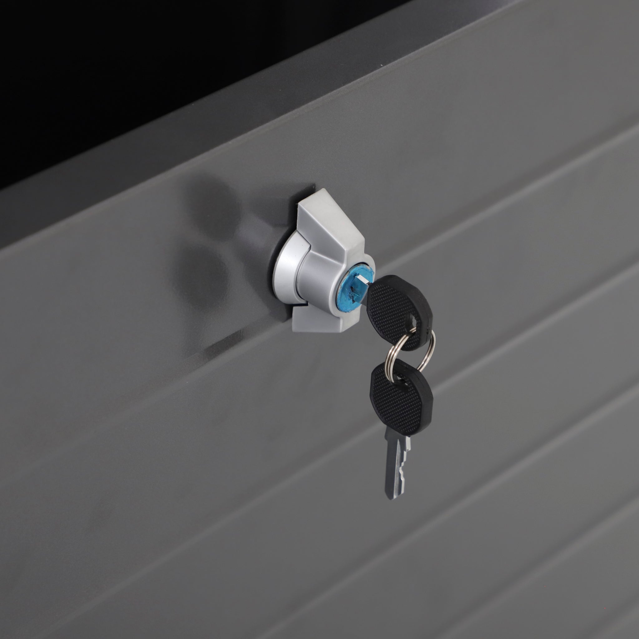 A close-up of a lock with two keys inserted in the black galvanised steel surface of a Pacific Lifestyle - Rhode Island Large Outdoor Cushion Box.