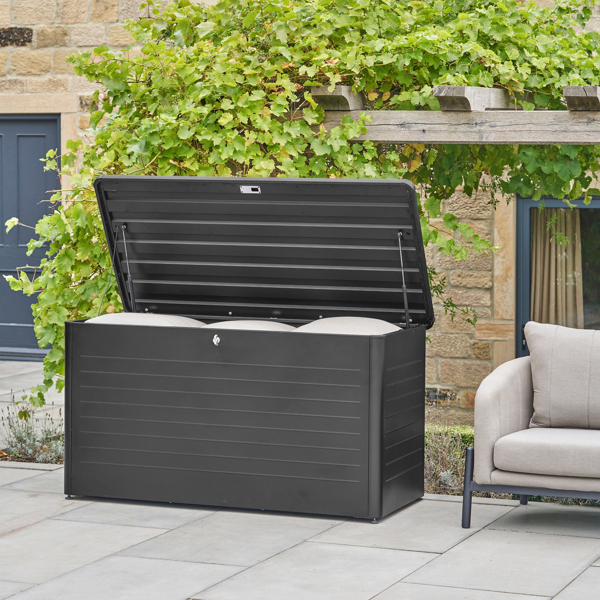 A Pacific Lifestyle Rhode Island Large Outdoor Cushion Box in black, shown with its lid open, is placed beside a patio sofa and surrounded by lush greenery.