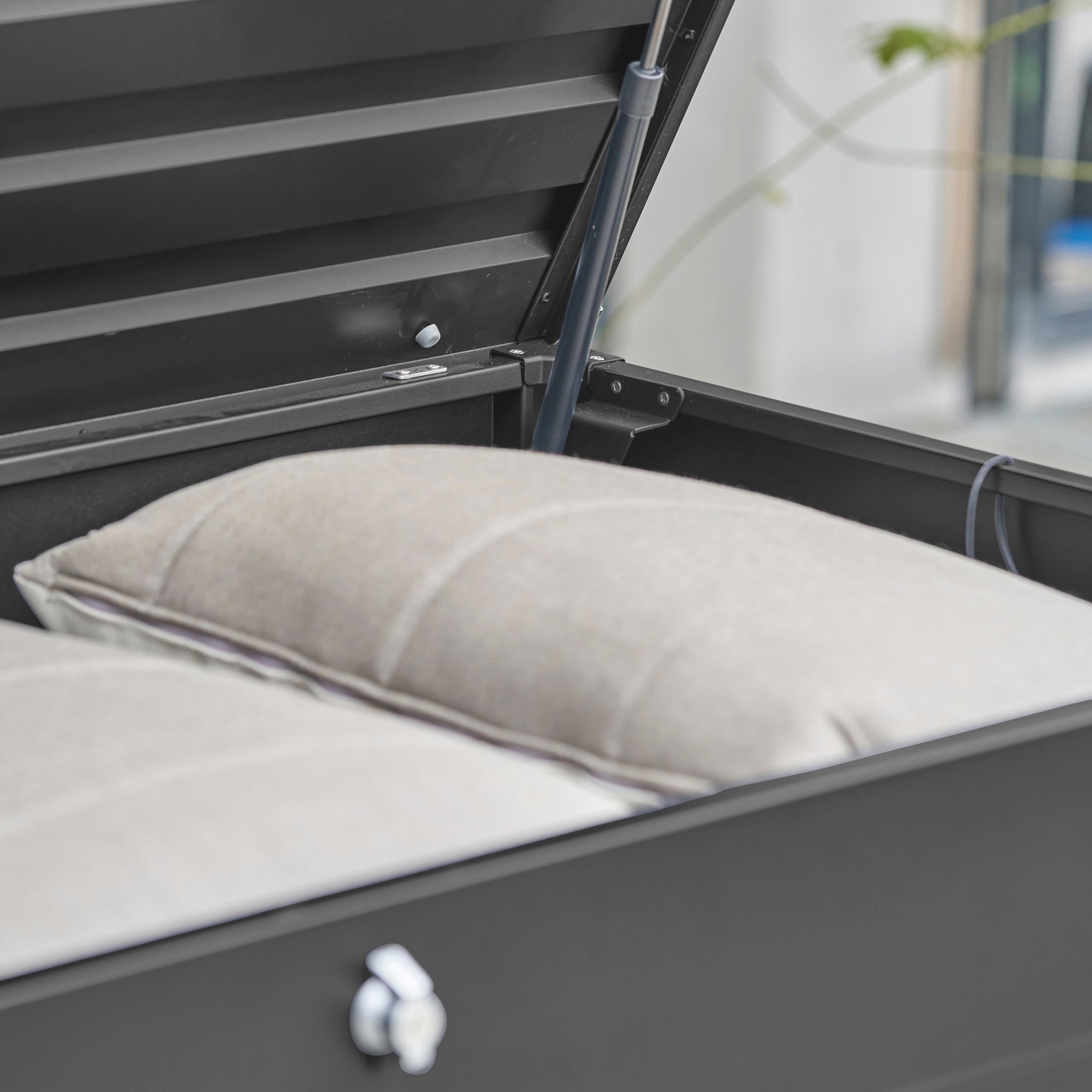 Close-up of a Pacific Lifestyle - Rhode Island Large Outdoor Cushion Box in Black, showing beige cushions inside and a metal hinge on the galvanised steel lid.