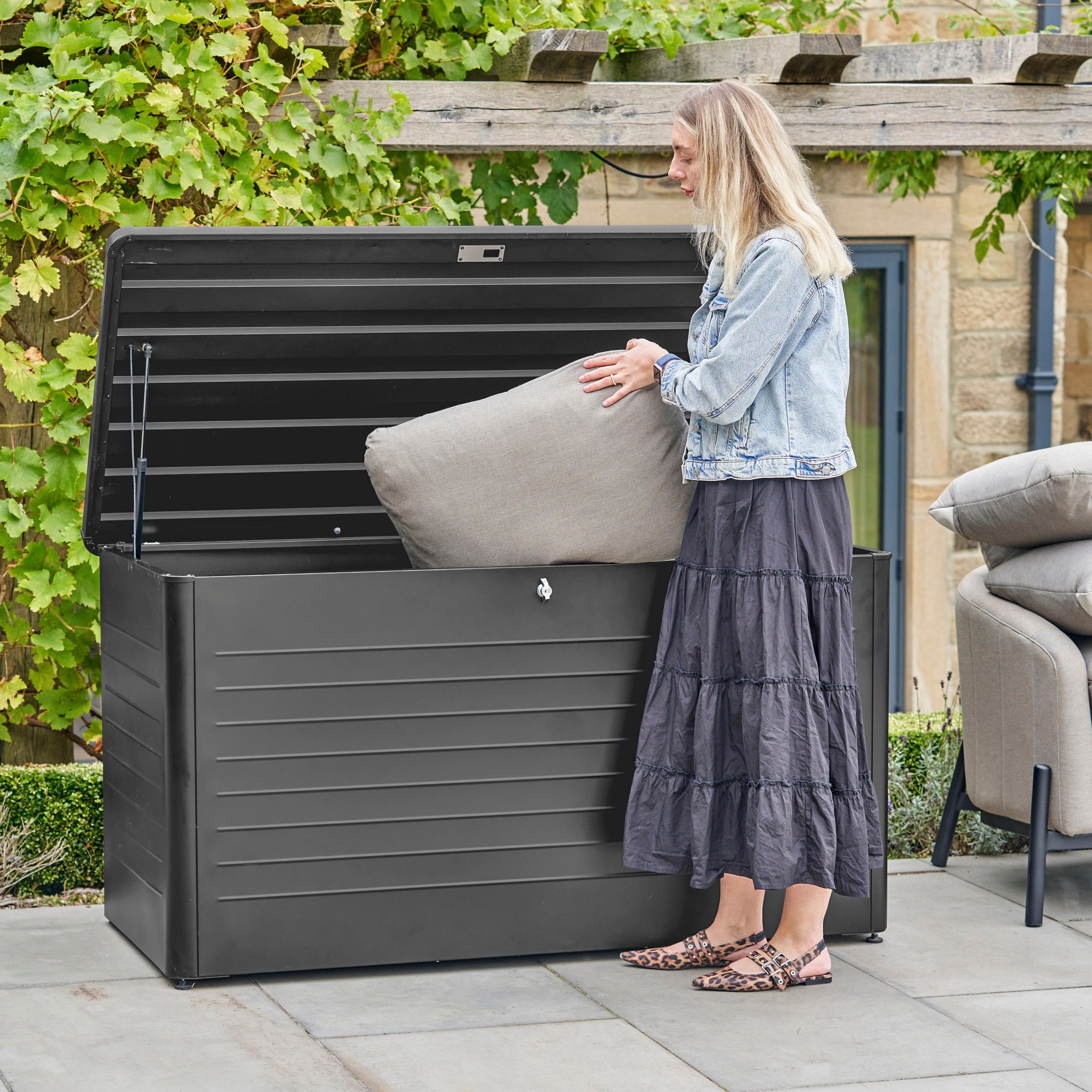A woman puts a cushion into the Pacific Lifestyle Rhode Island Large Outdoor Cushion Box in Black on a patio beside a vine-covered wall.