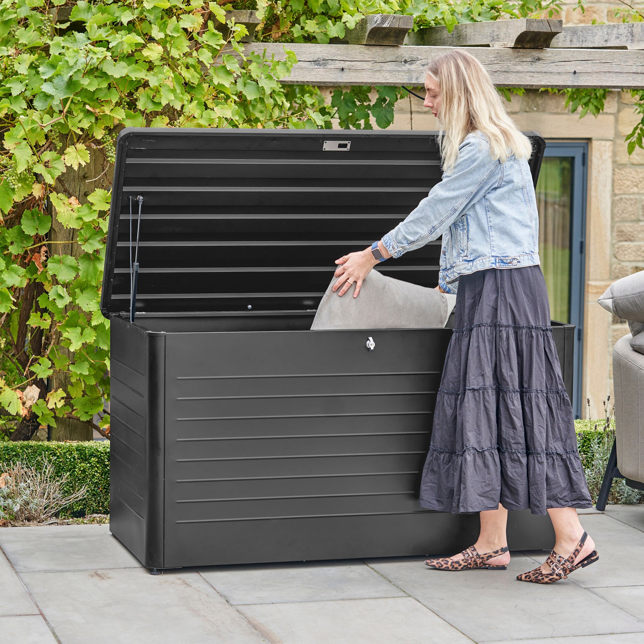 A woman places a cushion in the Pacific Lifestyle - Rhode Island Large Outdoor Cushion Box in Black on her patio. Made from durable galvanised steel, this box keeps patio essentials secure and protected from the elements.