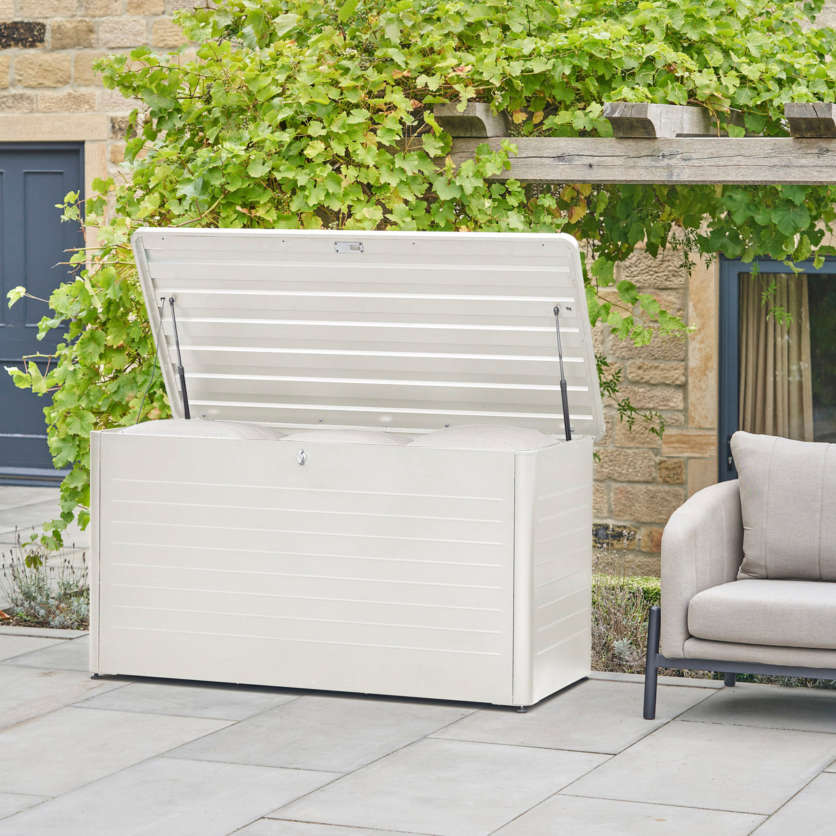 The Pacific Lifestyle Rhode Island Large Outdoor Cushion Box in Pebble, with its lid open, sits on a patio next to a chair.
