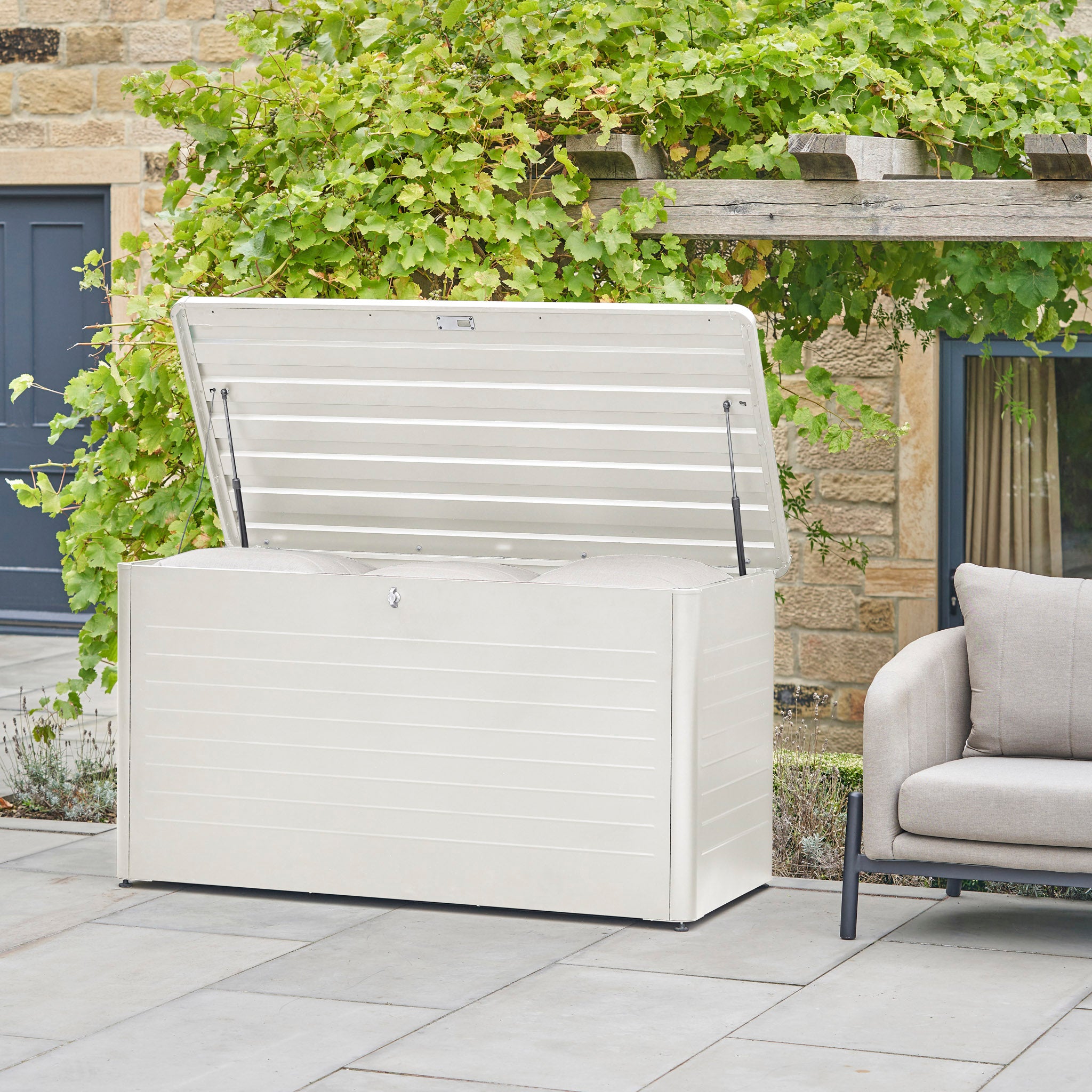 A Pacific Lifestyle - Rhode Island Large Outdoor Cushion Box in Pebble sits on a patio beside a gray cushioned chair, providing stylish and durable outdoor storage.