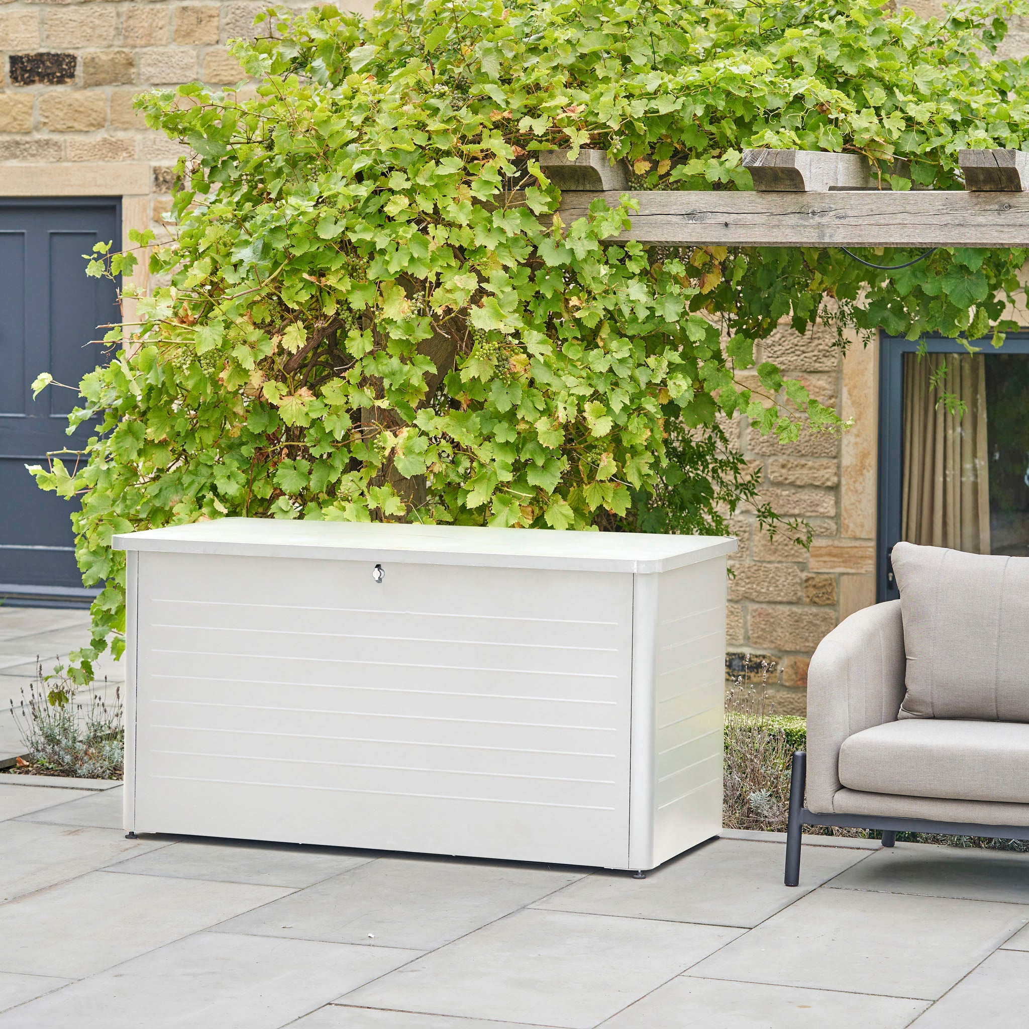 The Pacific Lifestyle - Rhode Island Large Outdoor Cushion Box in Pebble sits on a patio by a vine-covered pergola and beige cushioned chair, offering the perfect stylish storage for your garden essentials.