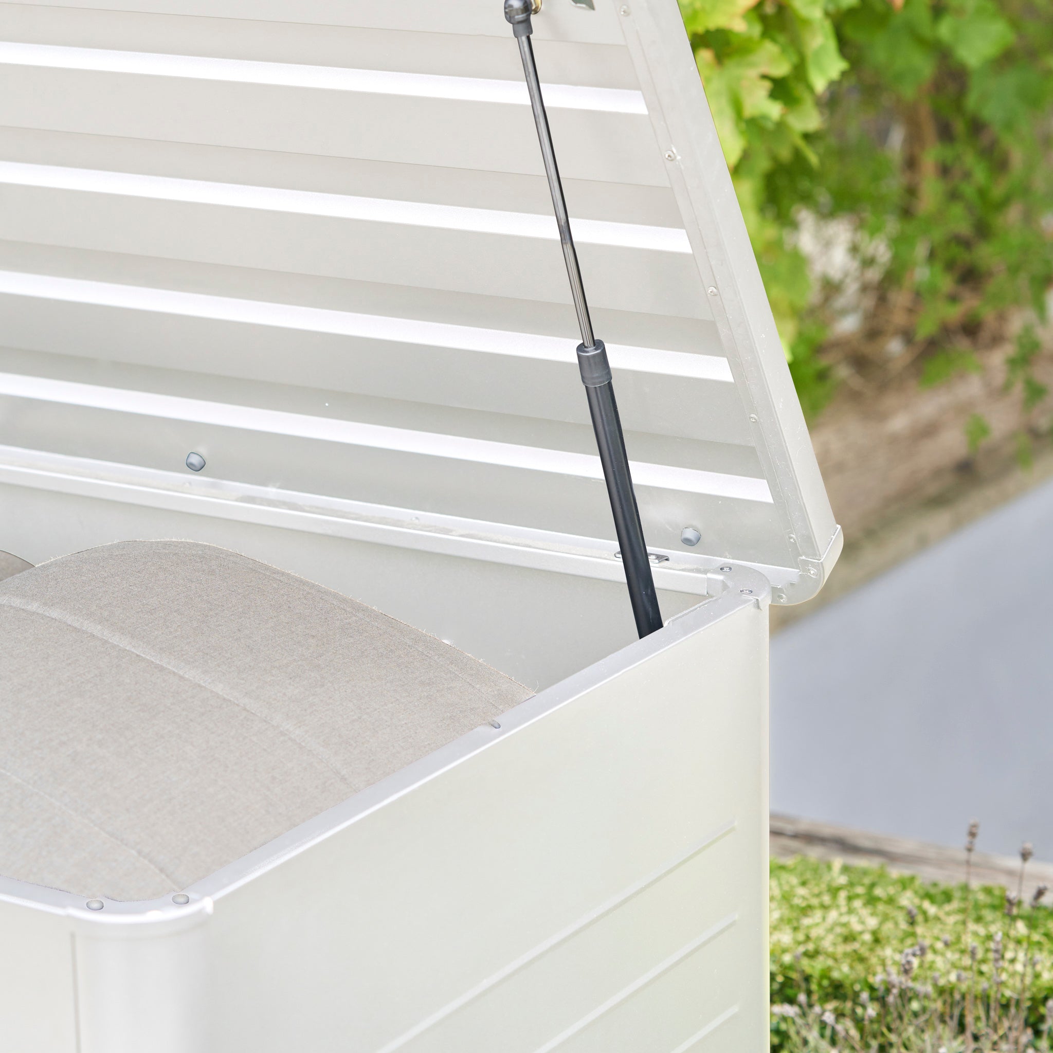Open the Pacific Lifestyle - Rhode Island Small Outdoor Cushion Box in Pebble, featuring a gas strut and beige cushions inside. Crafted from galvanized steel, this stylish storage box offers durable outdoor protection.