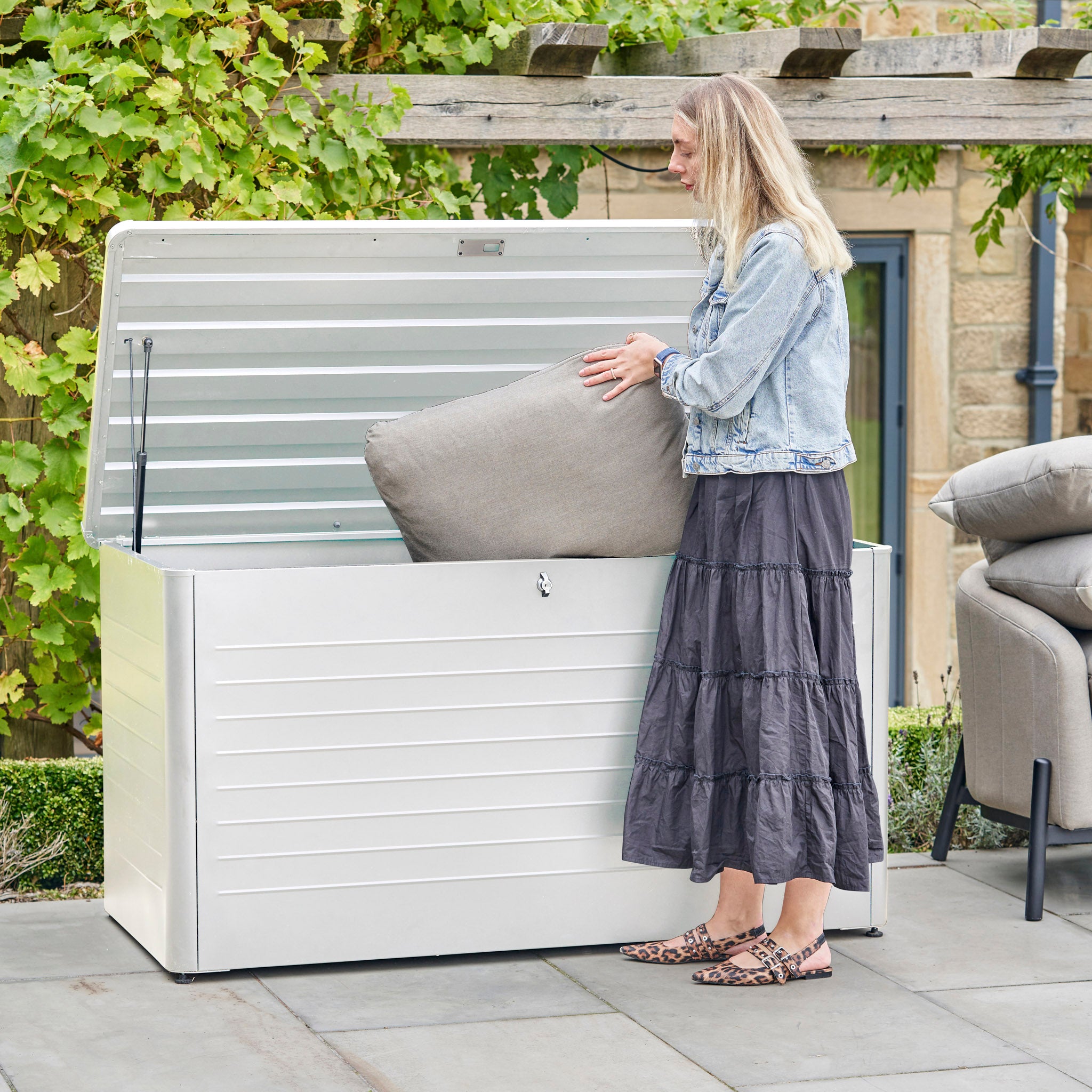 A woman places a cushion into the Pacific Lifestyle - Rhode Island Large Outdoor Cushion Box in Pebble on her patio.