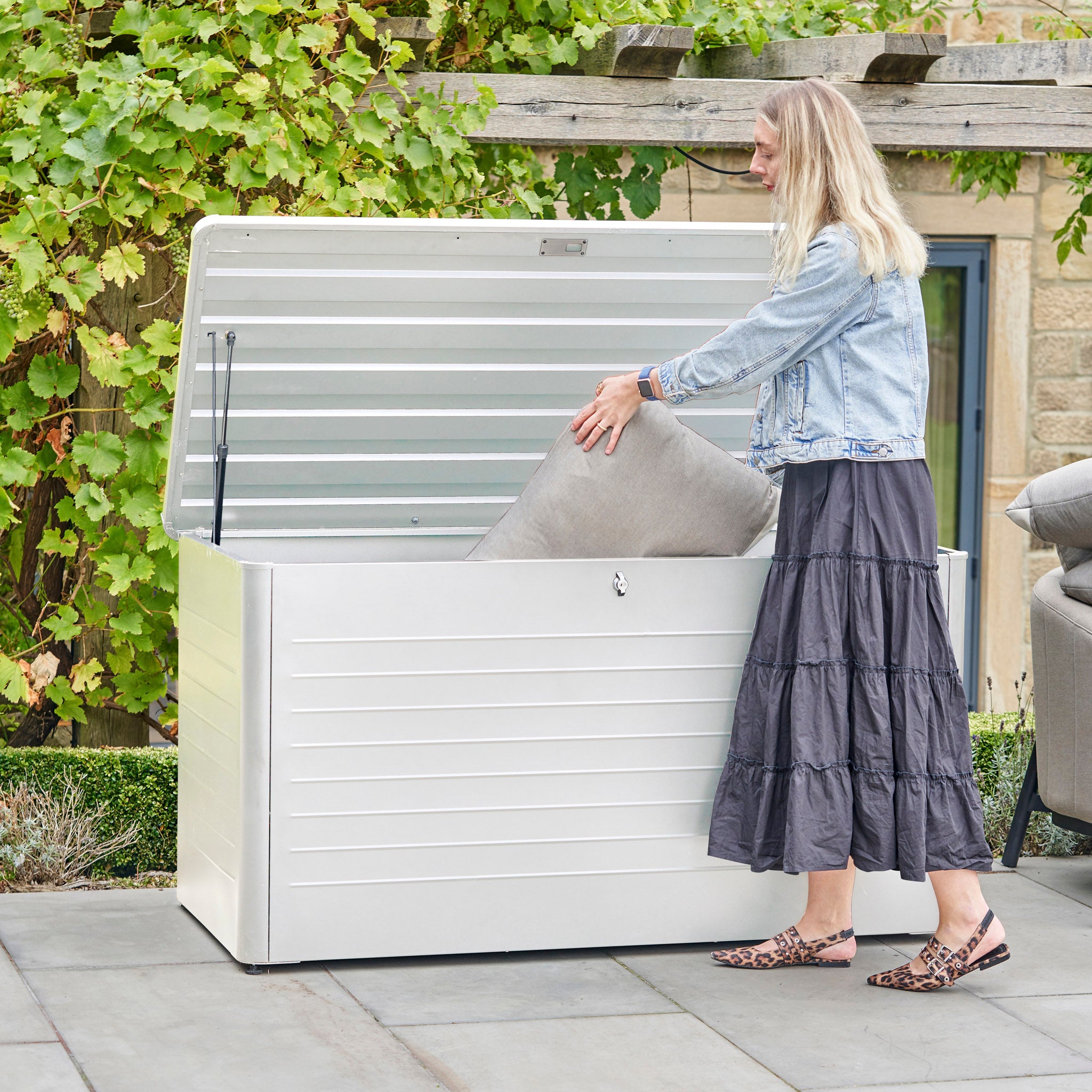A woman places a cushion into the Pacific Lifestyle - Rhode Island Large Outdoor Cushion Box in Pebble on a patio surrounded by plants. This durable galvanised steel box keeps cushions protected and your outdoor space tidy.