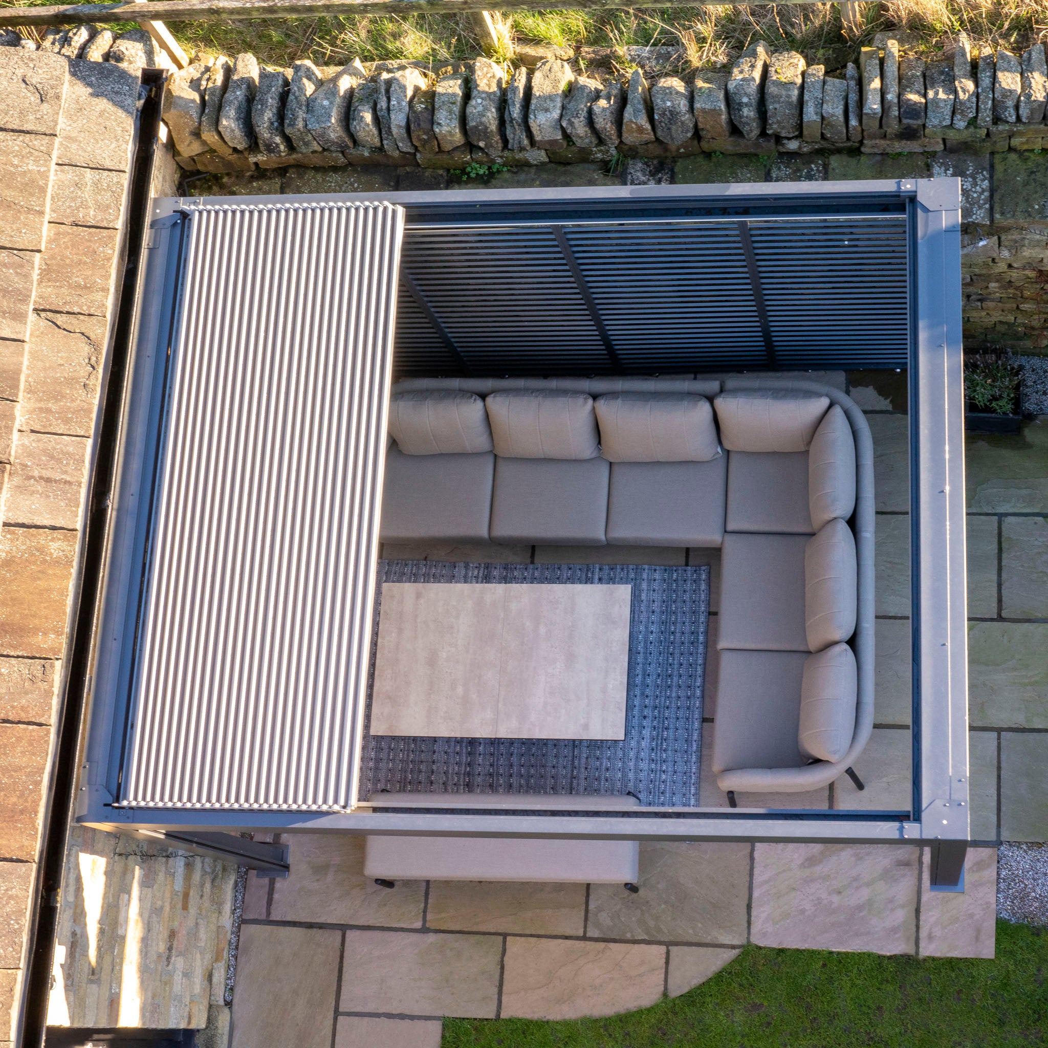 Aerial view of an outdoor space with a beige L-shaped sofa, table, and the Abierto Motorised 3m x 4m Pergola in grey featuring a retractable roof, LED lighting, and stone paving. Installation and delivery included.