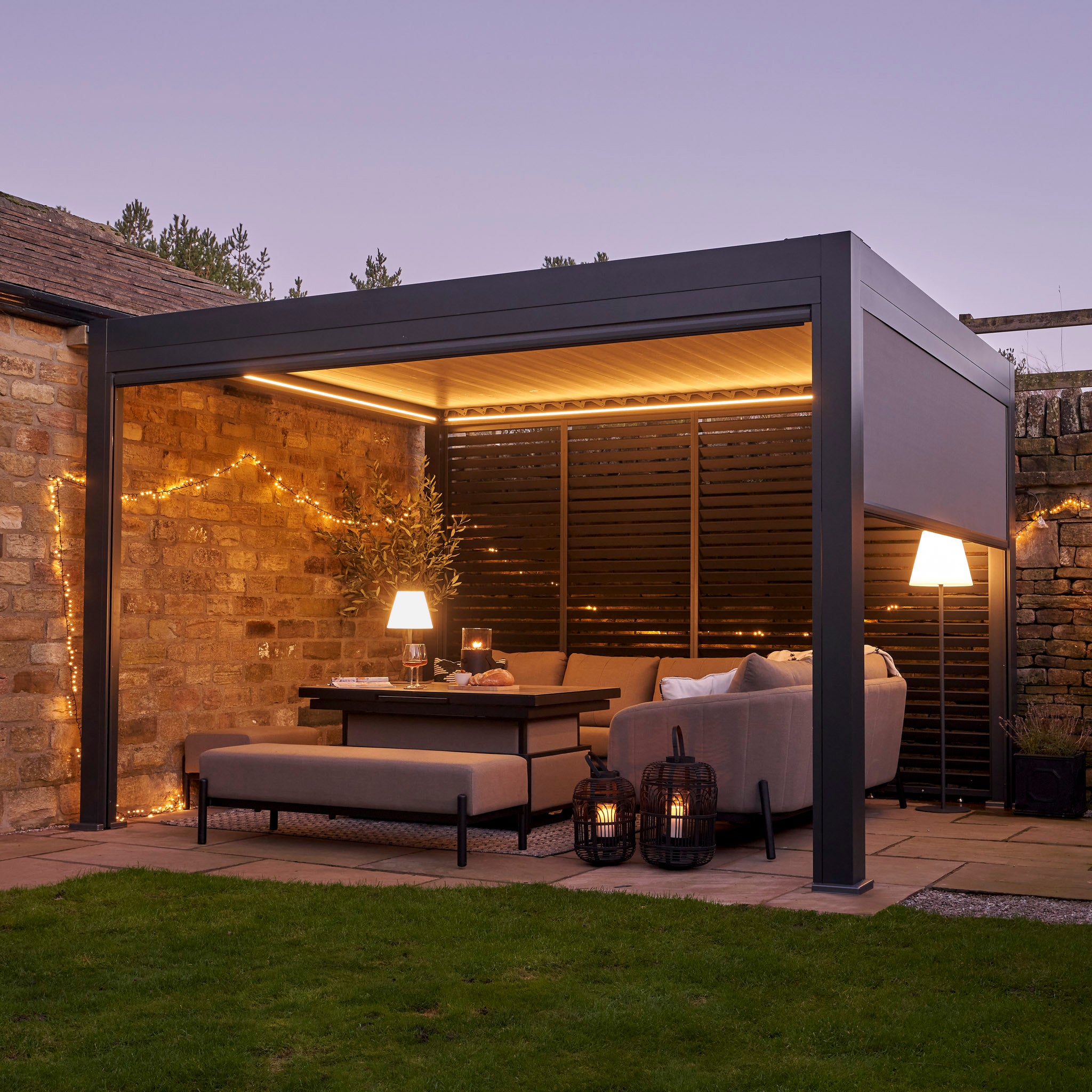The Abierto Motorised 3m x 4m Grey Pergola with retractable roof and LED lighting creates a modern outdoor lounge—perfect for relaxing on sofas beneath sleek illumination at dusk. Installation and delivery included.