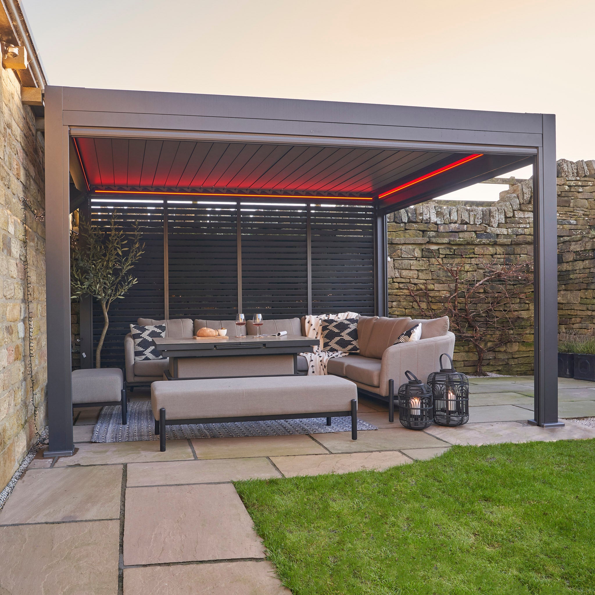 Create an inviting outdoor space with the Abierto Motorised 3m x 4m Pergola in grey featuring a retractable roof and integrated LED lighting—perfect for modern patios with sofas, tables, and benches. Installation and delivery included.