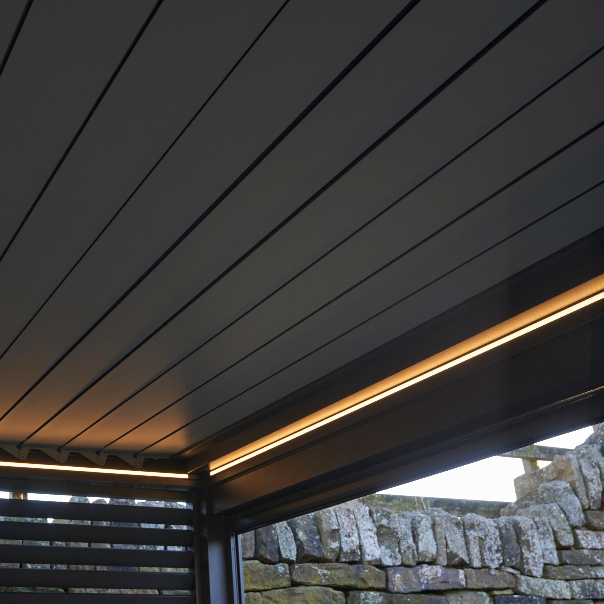 The Abierto Motorised 3m x 4m Pergola in grey features a retractable roof and built-in LED lighting, installed beside a stone wall with an outdoor view, creating a modern, stylish setting.