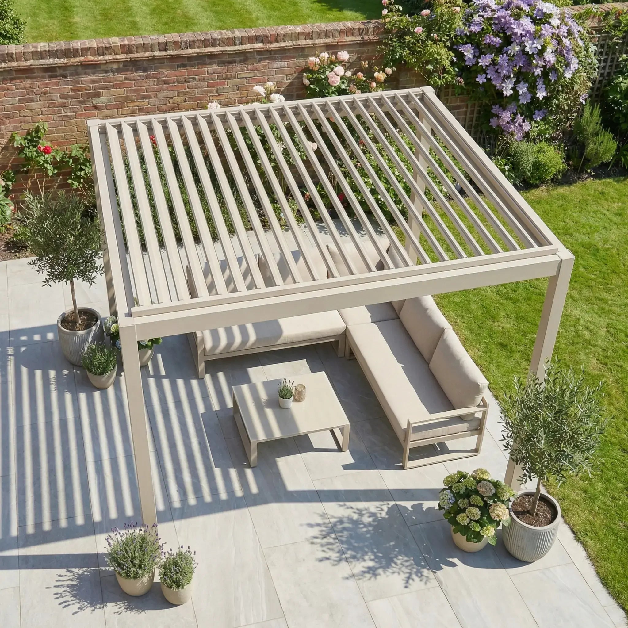 The PergoSTET Lite 2.4m x 3m Metal Rectangular Pergola in Latte with tilting louvres shades a patio furnished with a sectional sofa, table, and potted plants, set amid lush greenery.