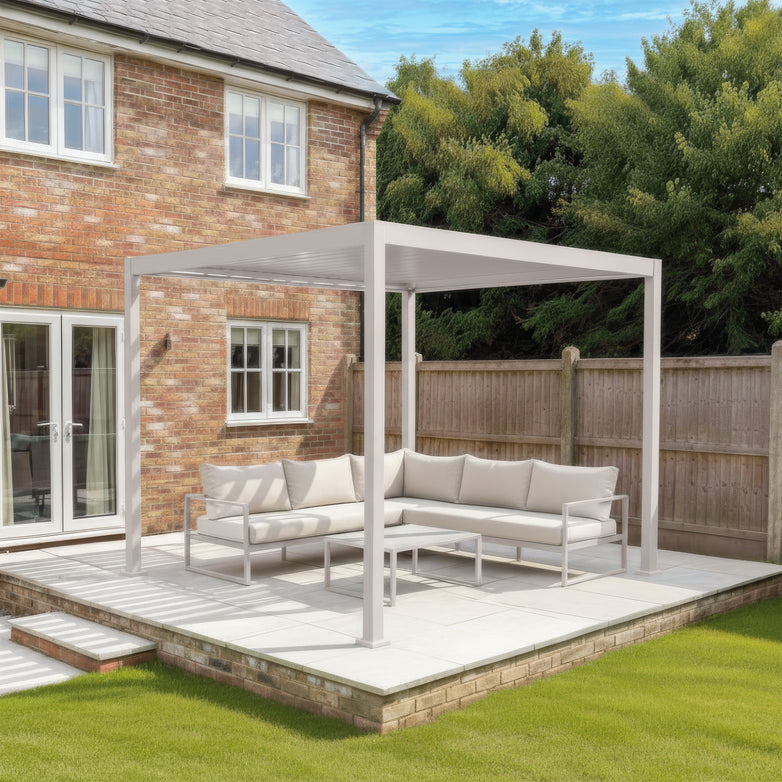 The PergoSTET Lite 2.4m x 3m Metal Rectangular Pergola in Latte features tilting louvres and cushioned seating, shown on a patio next to a brick house with lawn and trees in the background.