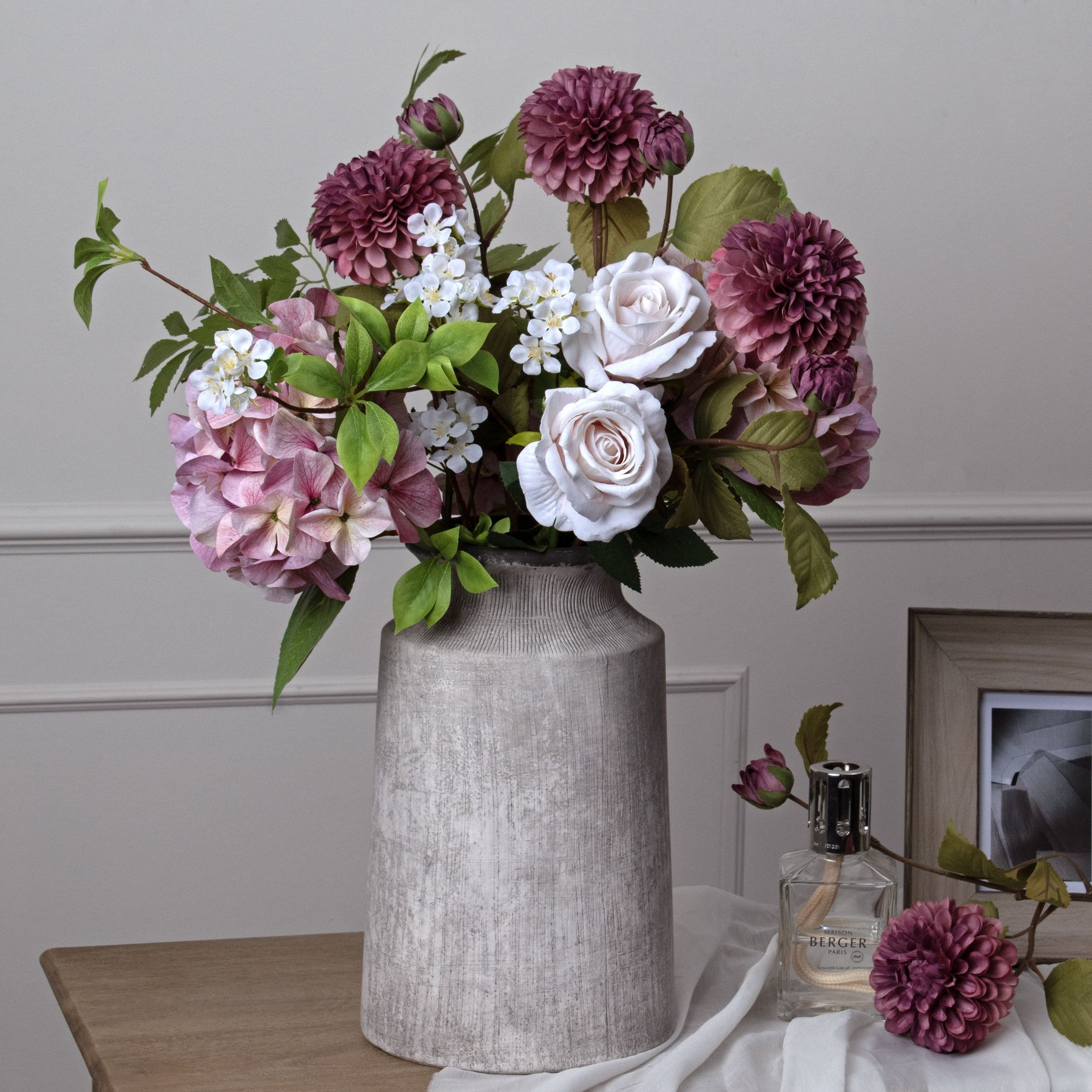 A gray ceramic vase holding pink and white flowers adorns a wooden table. It sits beside a framed photo, a fallen flower, and a perfume bottle. This Bloomville Stone Vase adds an elegant touch to your home décor with its subtle natural stone finish.