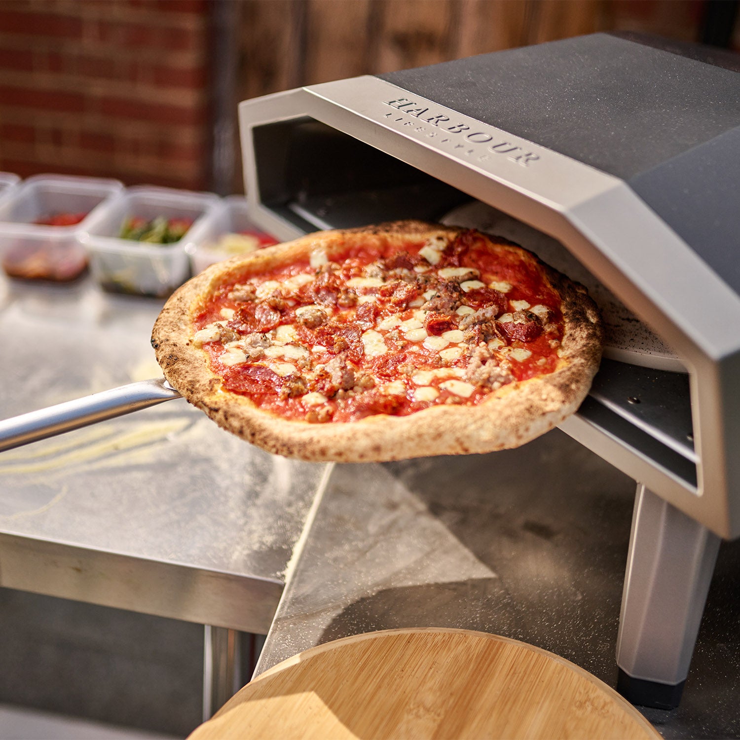 A freshly baked pizza with cheese and meat being pulled out of the Juno 16" Pizza Oven Bundle, using the included stainless steel pizza peel, from a portable outdoor oven with foldable legs.