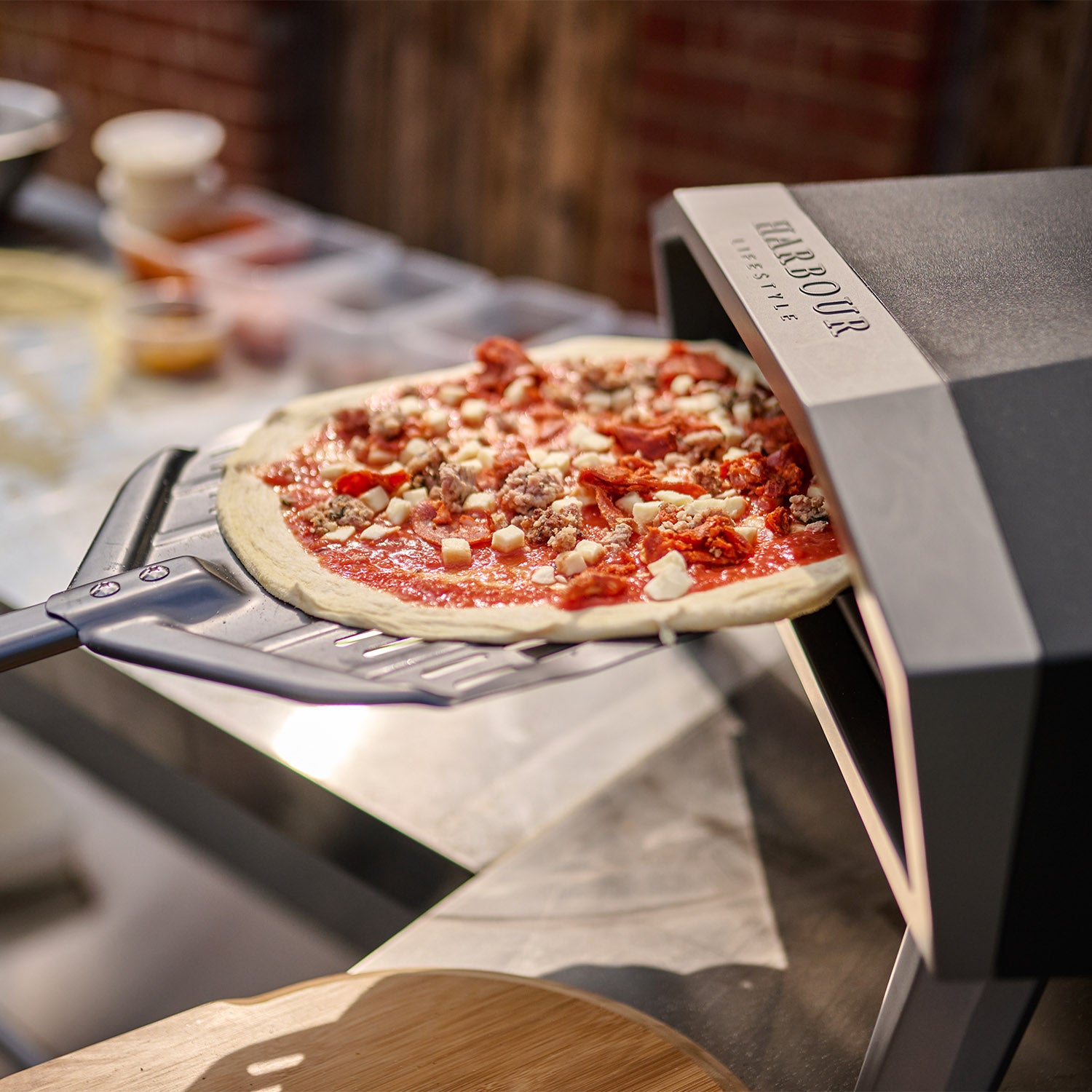 A pizza on a stainless steel peel is being placed into a Juno 16" Pizza Oven Bundle with foldable legs on a patio. Ingredients are visible in the background, while the built-in heat gauge thermometer ensures perfect cooking.