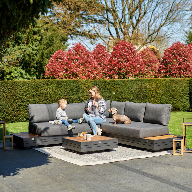 A woman and child sit on a Panama Luxury Outdoor Corner Group Set in Charcoal with a dog on a sunny day, surrounded by greenery and red bushes.