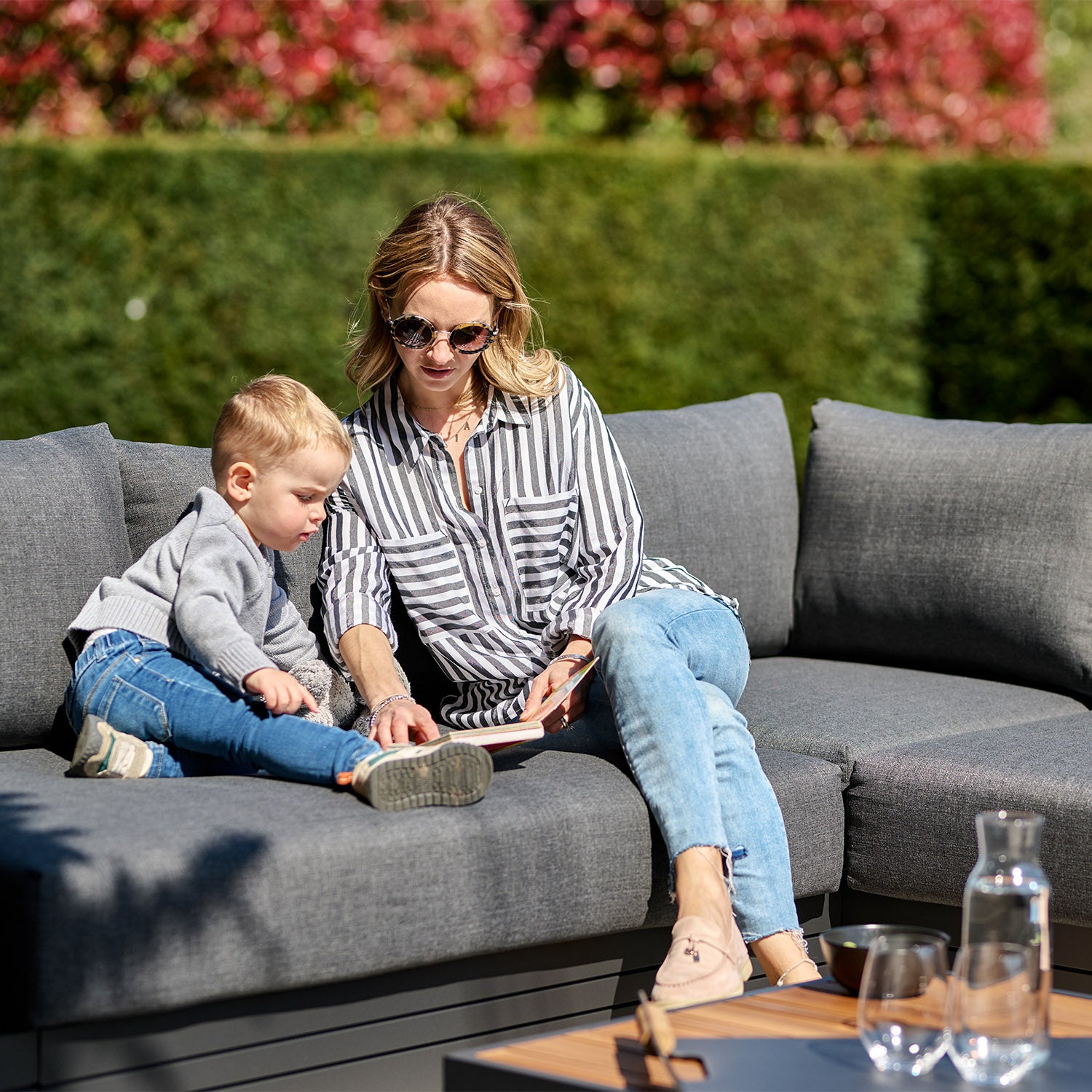 A woman and a child sit on the Panama Luxury Outdoor Corner Group Set in Charcoal, looking at a tablet together on a sunny day with green hedges in the background.