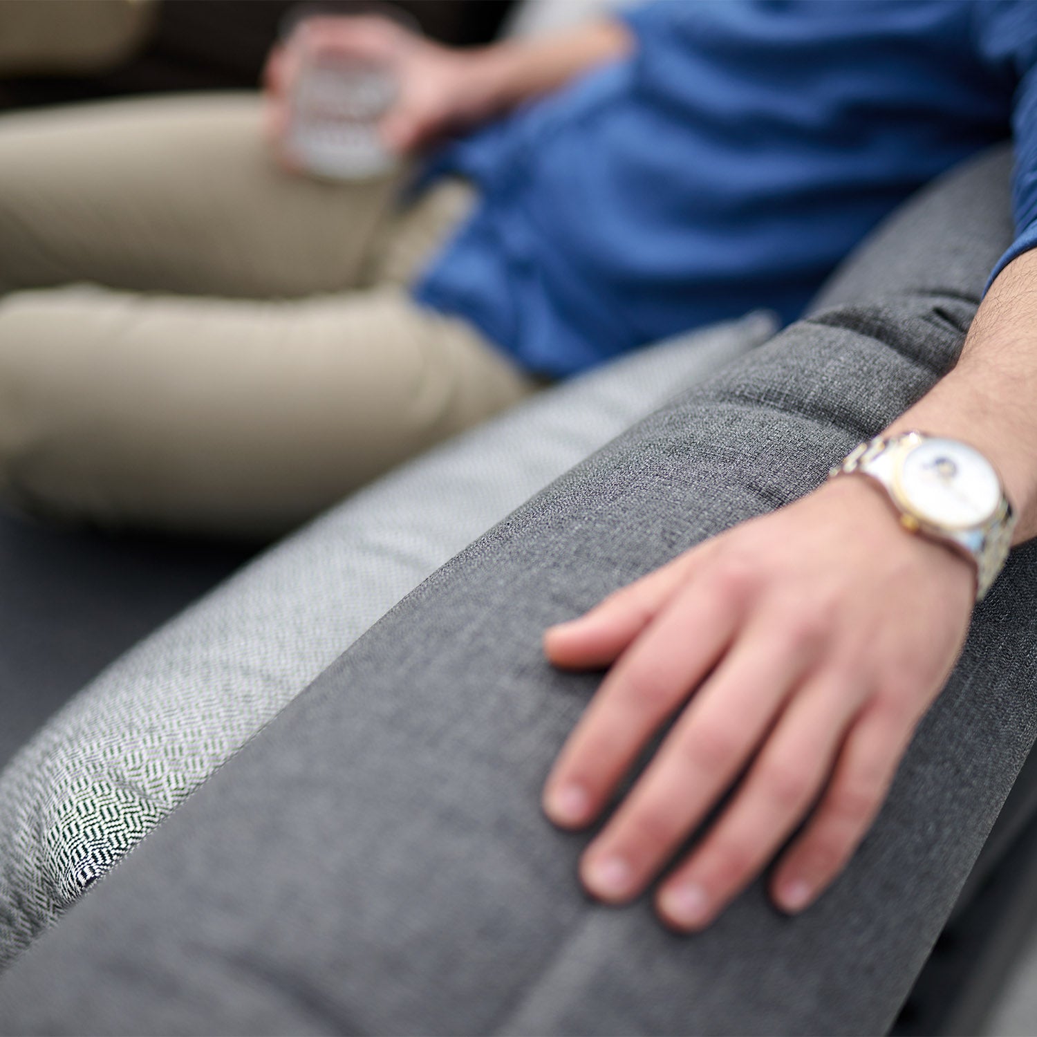 A person in a blue shirt relaxes on the Panama Luxury Outdoor Corner Group Set in Charcoal, holding a drink, with their hand resting on the armrest.