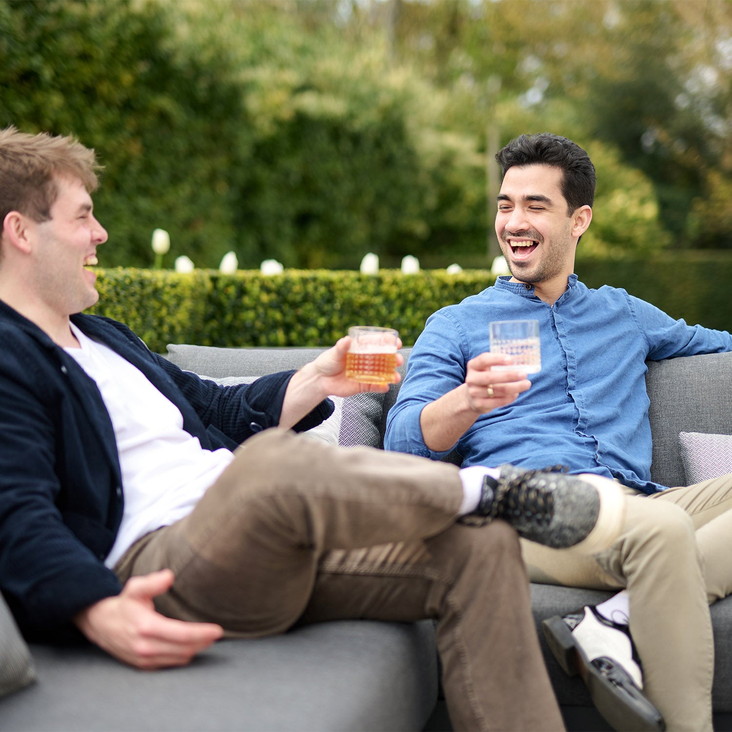 Two men sitting on a Panama Luxury Outdoor Corner Group Set in Charcoal, smiling and holding drinks, enjoying a casual conversation.