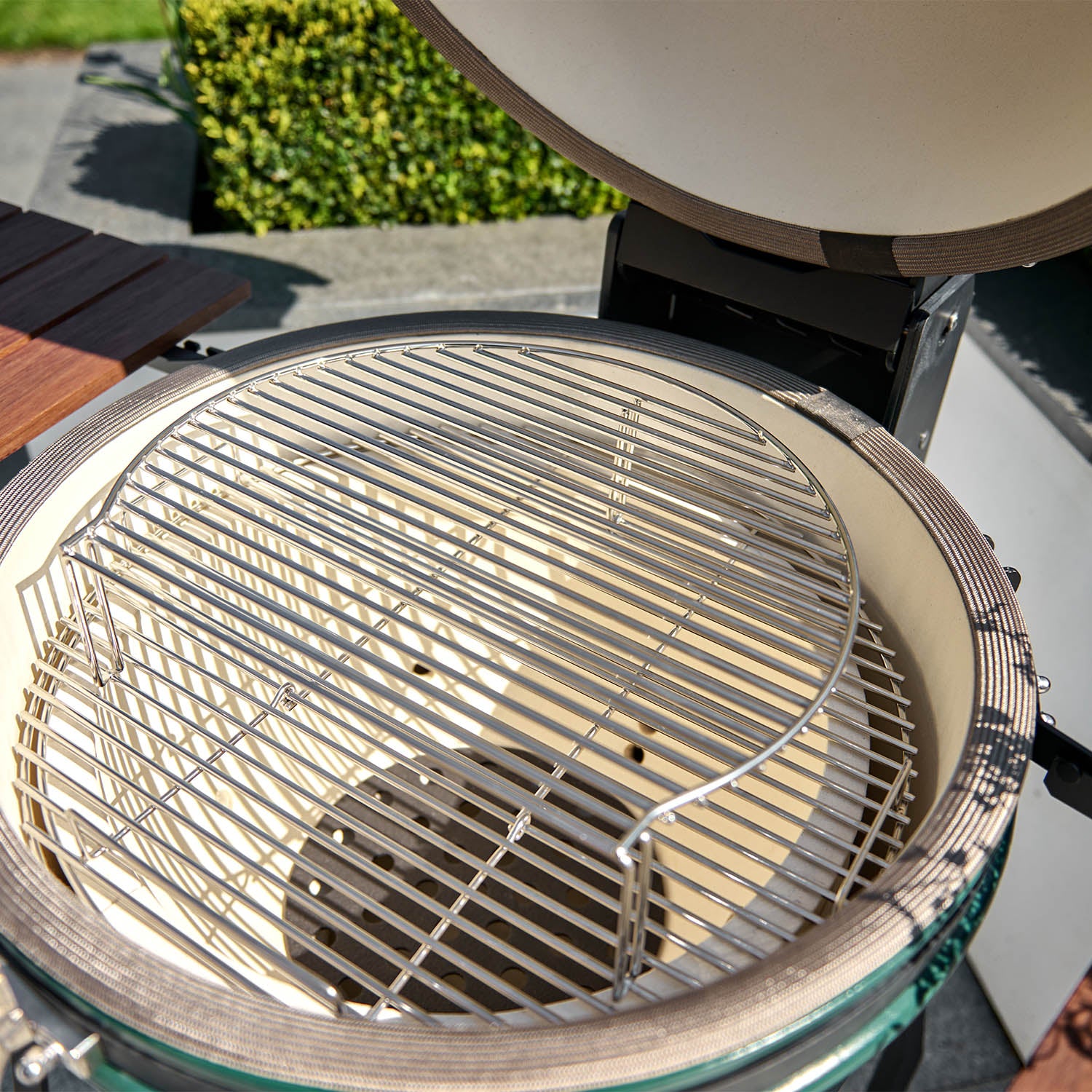 The outdoor ceramic Kamado Grill, with its open lid showcasing a clean, circular metal grilling grate, features the Cooking Grid Expander for 18" Kamado Grills which offers 60% more cooking space. Green bushes can be seen in the background.