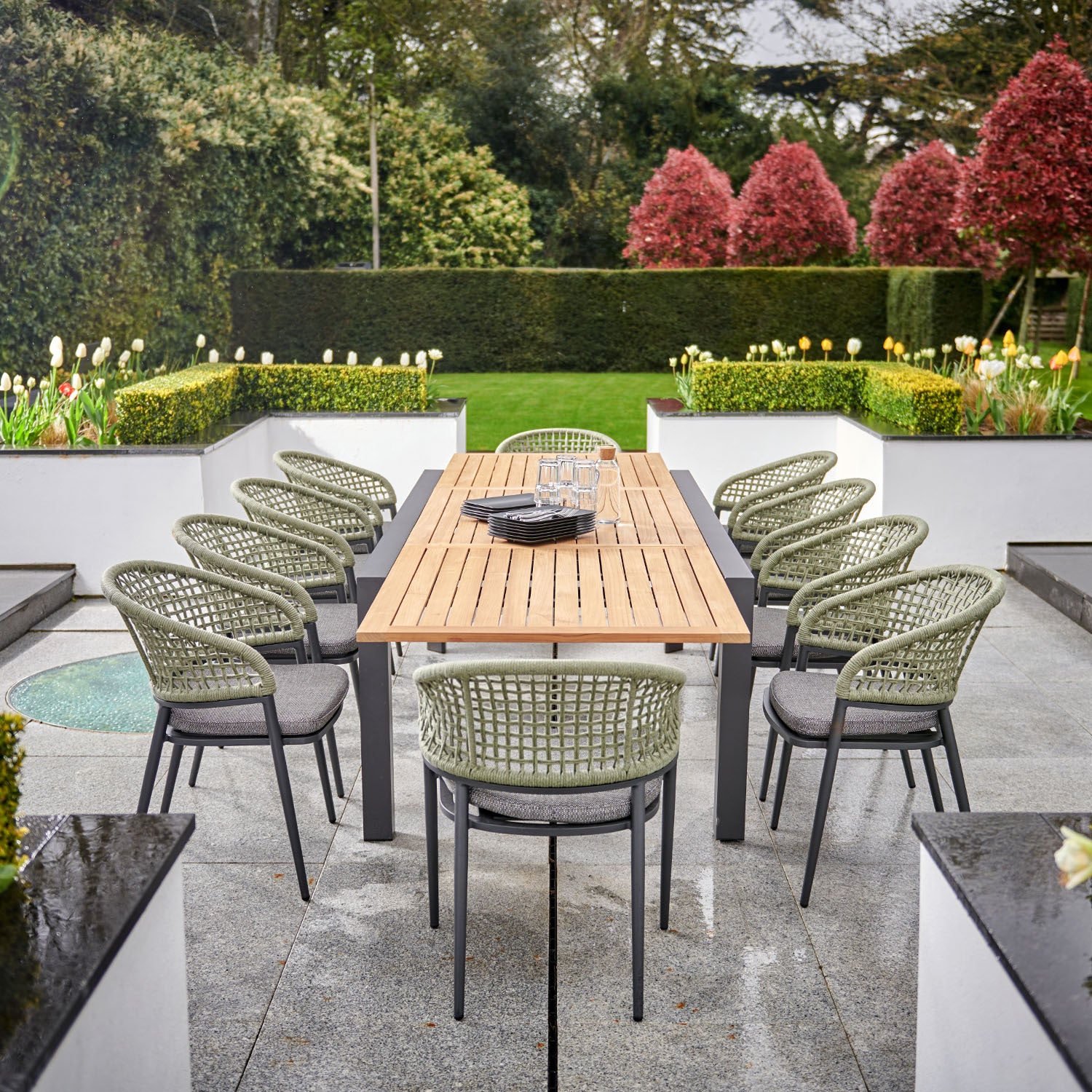 Outdoor dining area featuring the Kalama 10 Seat Rectangular Extending Dining Set with a teak table in olive green, surrounded by woven chairs and set on a stone patio with garden views.