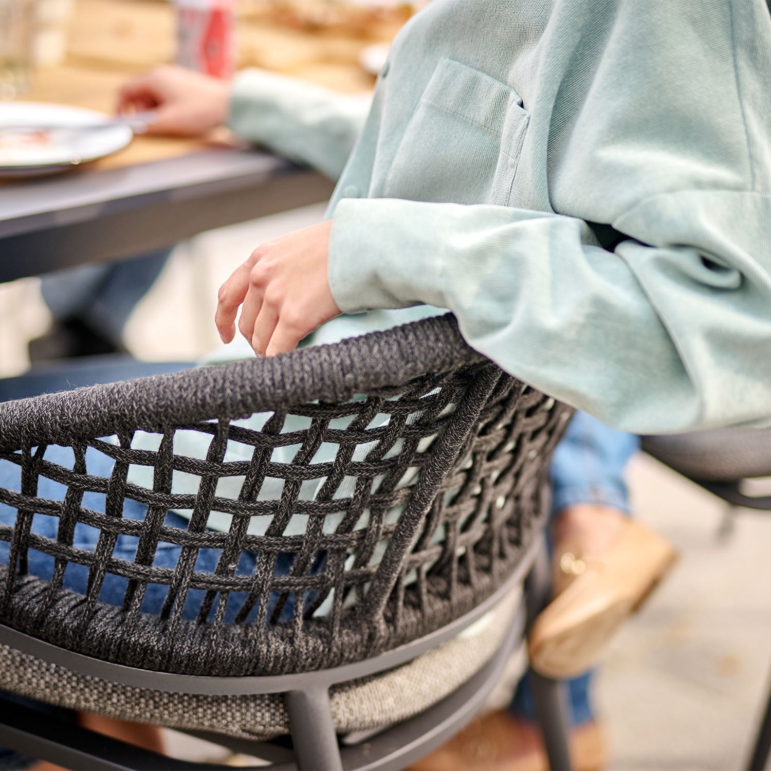 A person in a green sweater is sitting on one of the chairs from the Kalama 8 Seat Rectangular Dining Set with Teak Table in Charcoal, surrounded by a blurred background.