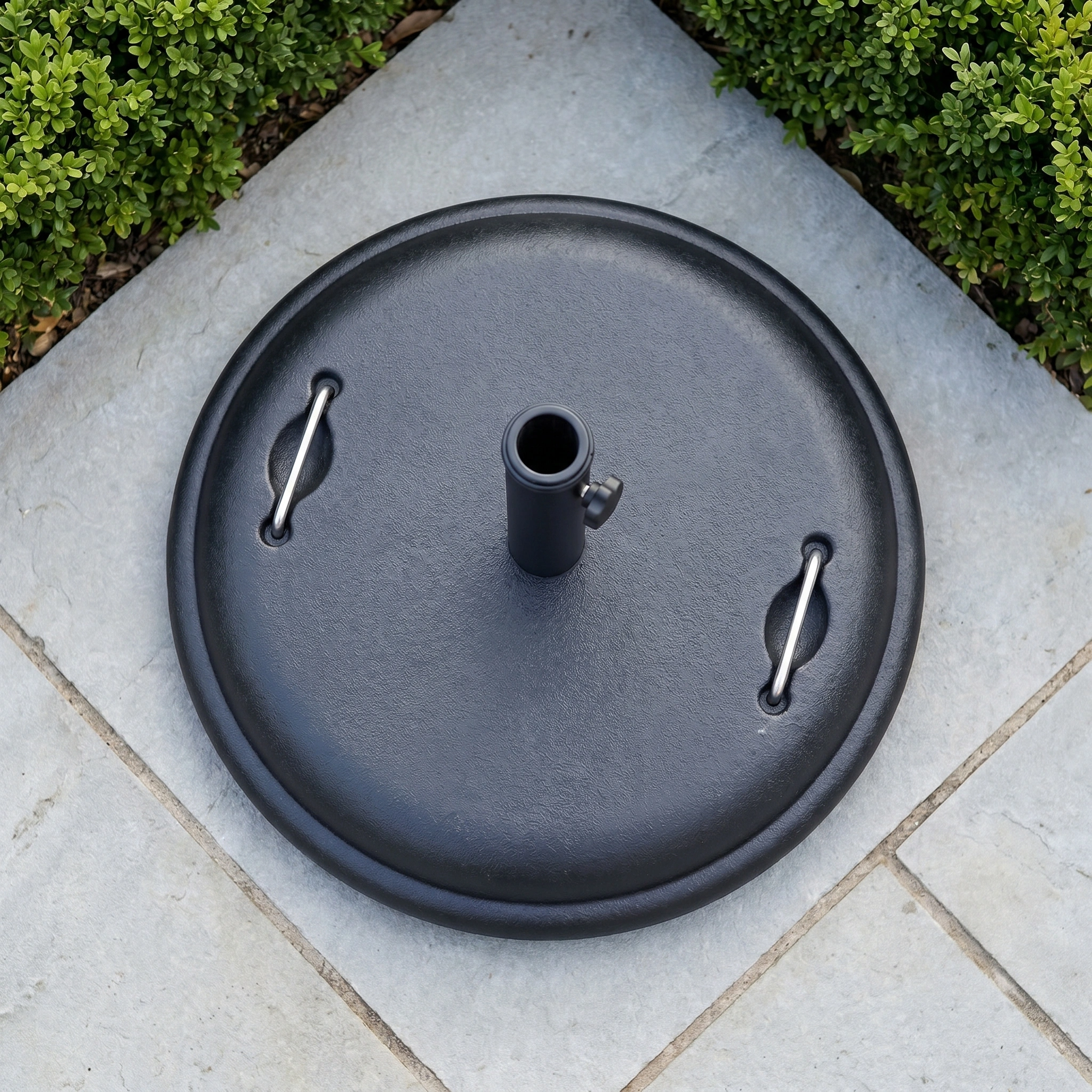 The 25kg Concrete Parasol Base sits on stone patio tiles, surrounded by neatly trimmed green bushes.