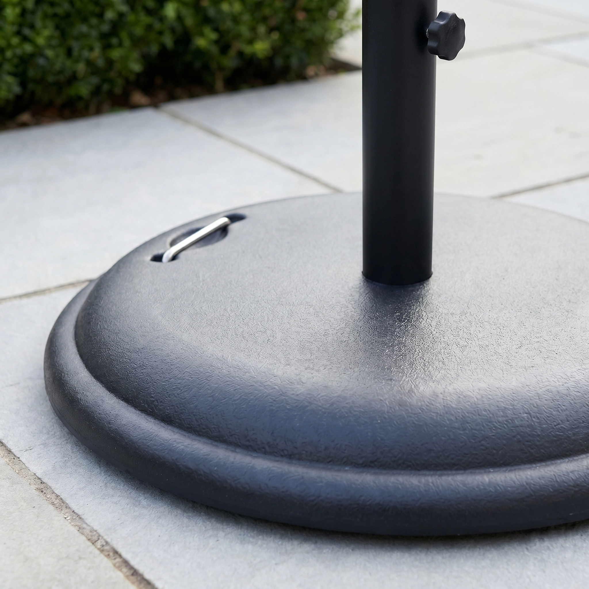 Close-up of the 25kg Concrete Parasol Base on a tiled patio, surrounded by lush greenery. The sturdy black base blends seamlessly with the modern outdoor setting.