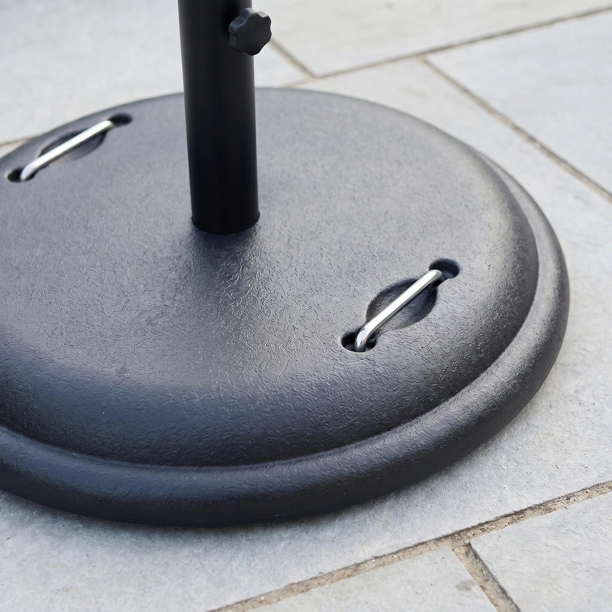 Close-up of the 25kg Concrete Parasol Base with metal loops on a tiled patio, highlighting its sturdy black concrete design for outdoor use.