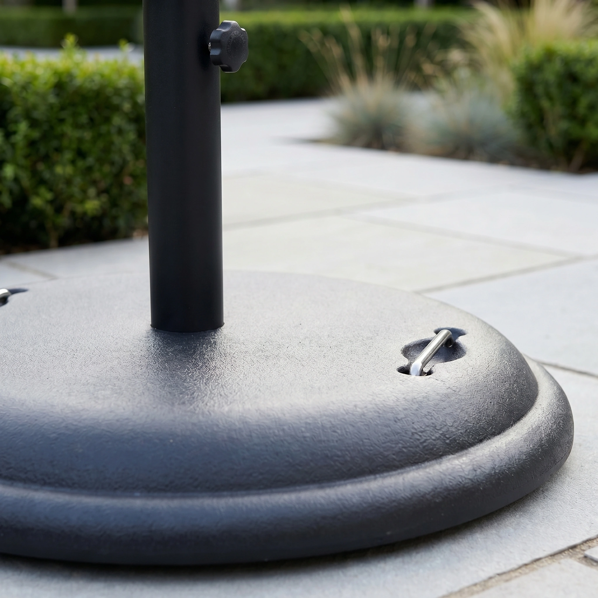 A close-up of the 25kg Concrete Parasol Base on a tiled patio, with green bushes in the background.