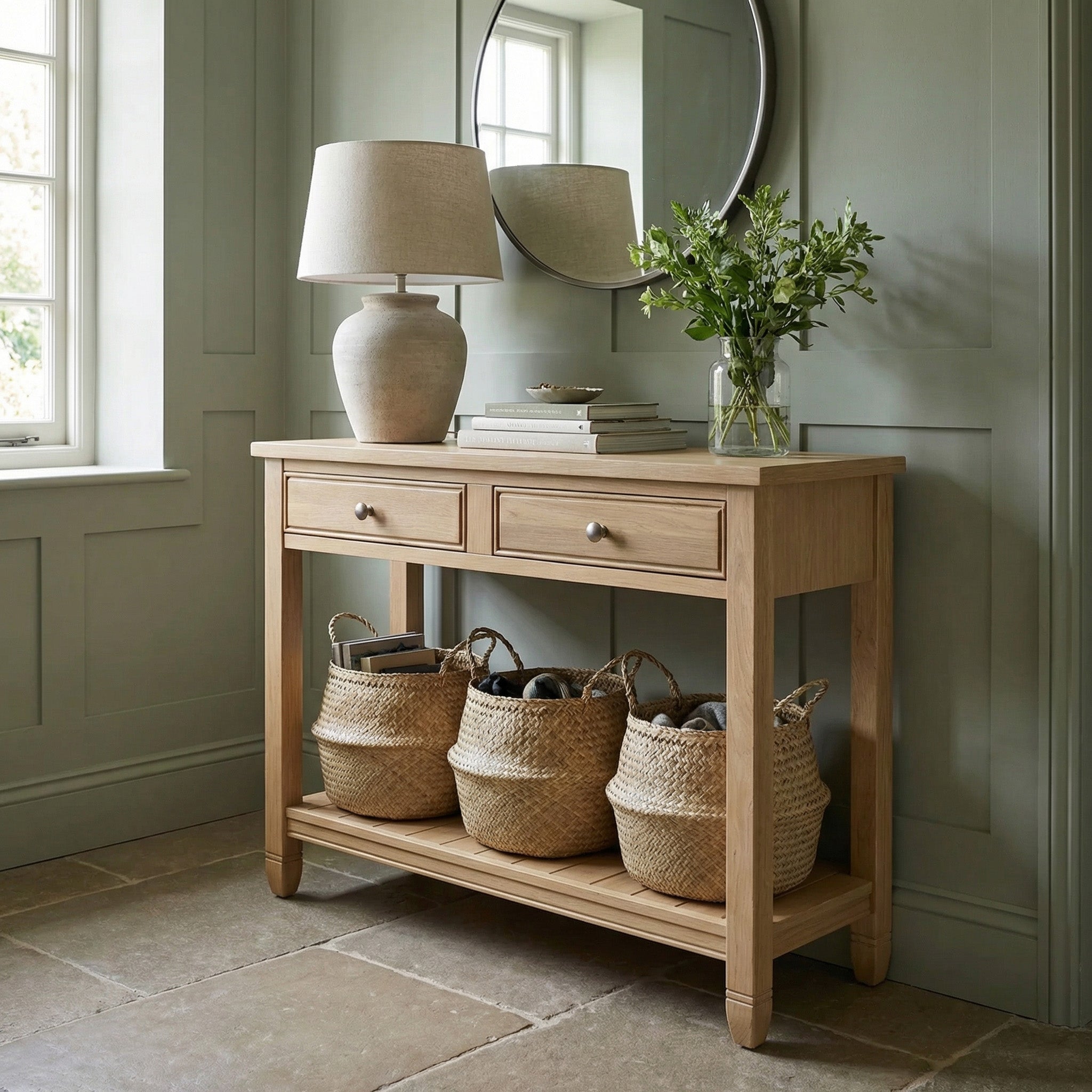 A light, airy hallway features the Amberley 2 Drawers Console Table in Light Oak, topped with a lamp and vase of flowers, with baskets underneath for both elegance and practical storage.
