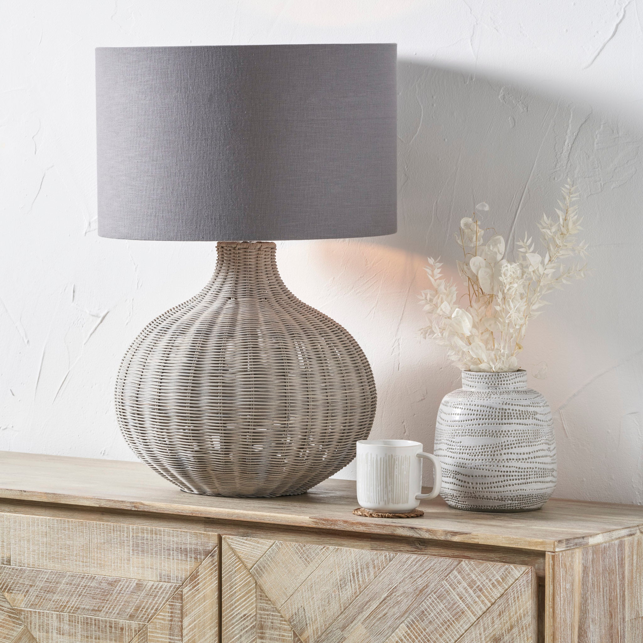 The Pacific Lifestyle Allona Grey Wash Rattan Table Lamp, featuring an organic tactile finish and gray shade, pairs beautifully with a wooden cabinet alongside a mug and dried flowers in a vase.