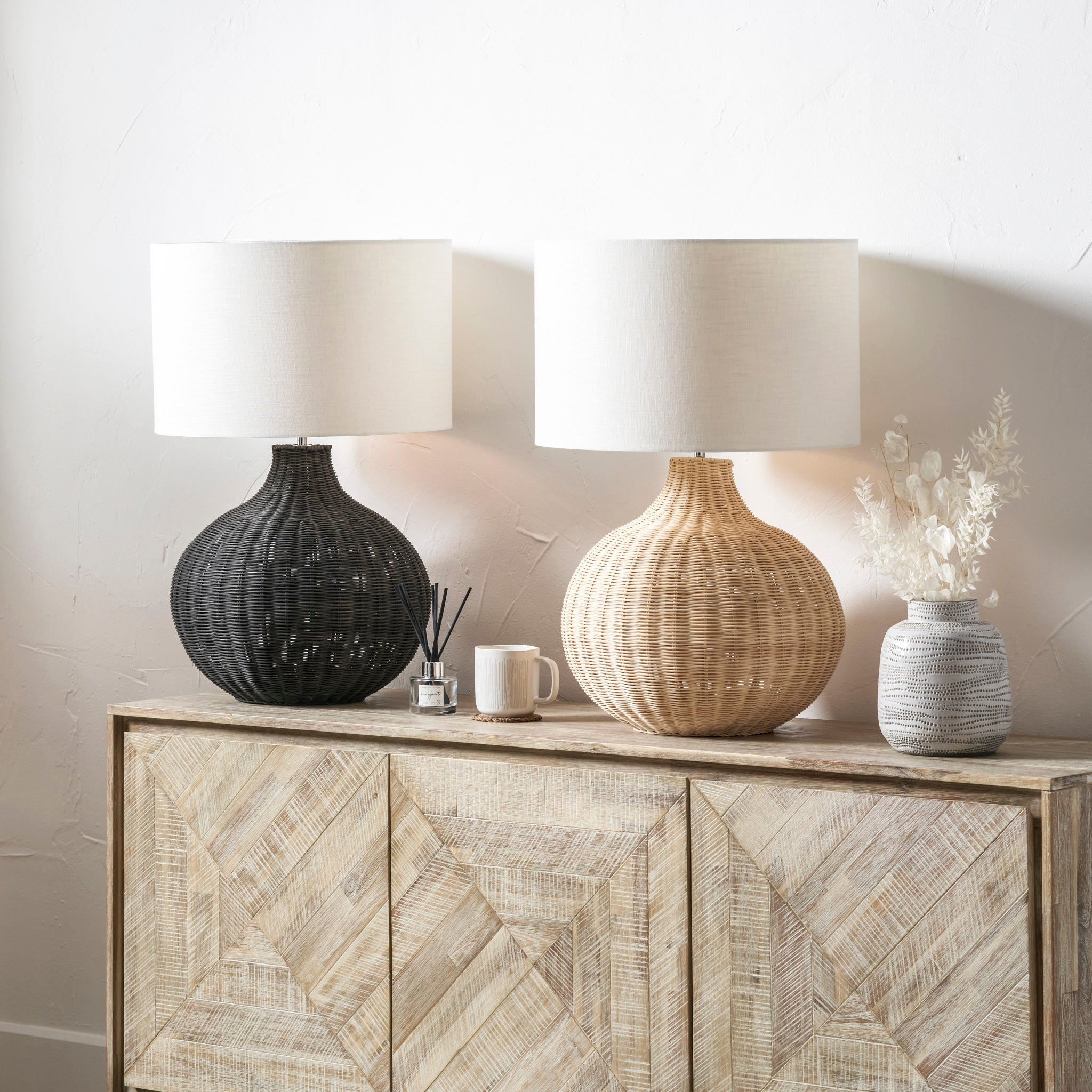 Two Allona Grey Wash Rattan Table Lamps from Pacific Lifestyle with white shades sit elegantly on a wooden sideboard; a small vase with dried flowers rests on the right, adding an organic tactile finish to the setting.