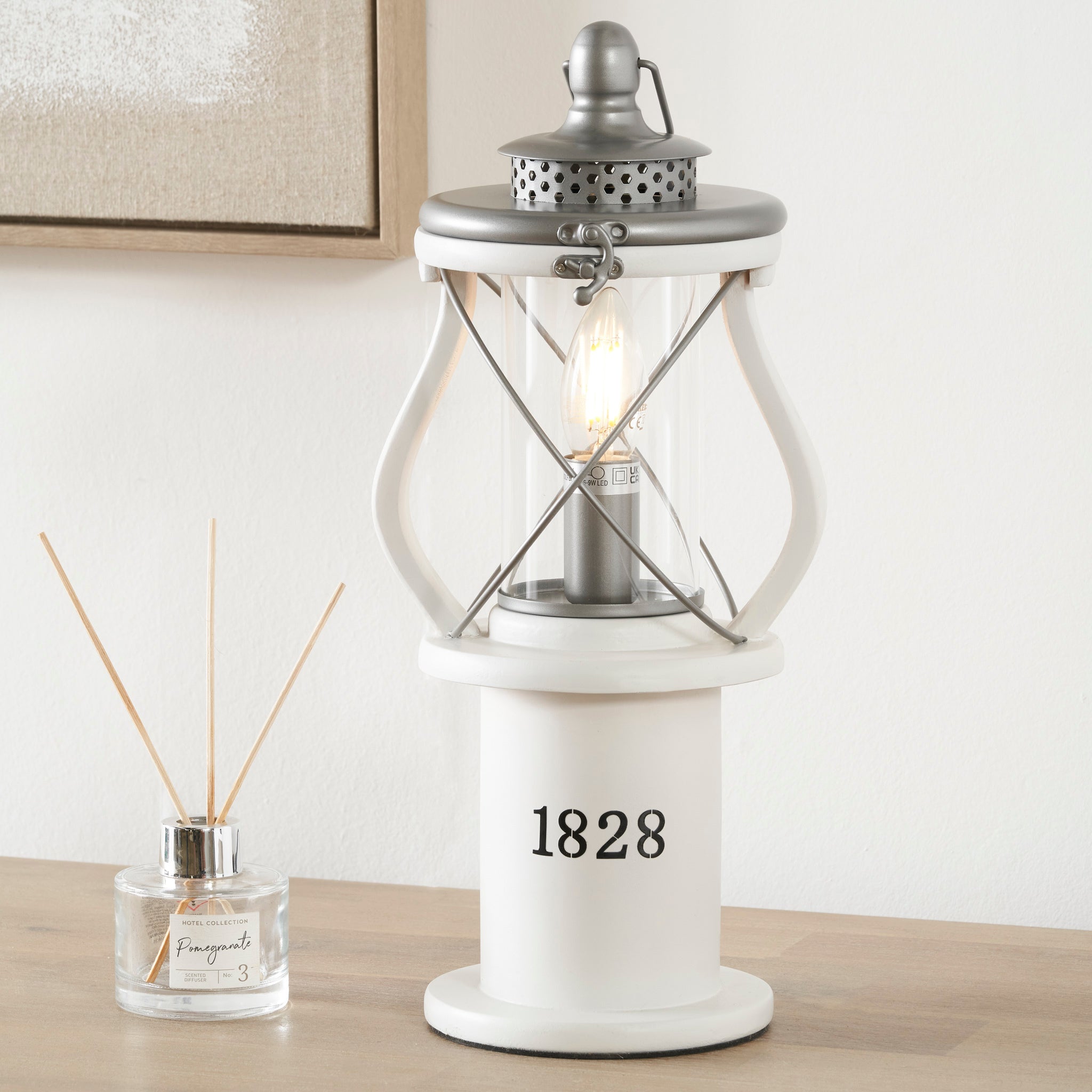 The Pacific Lifestyle Gibson Wood Lantern Table Lamp in White sits on a wooden surface next to a reed diffuser.