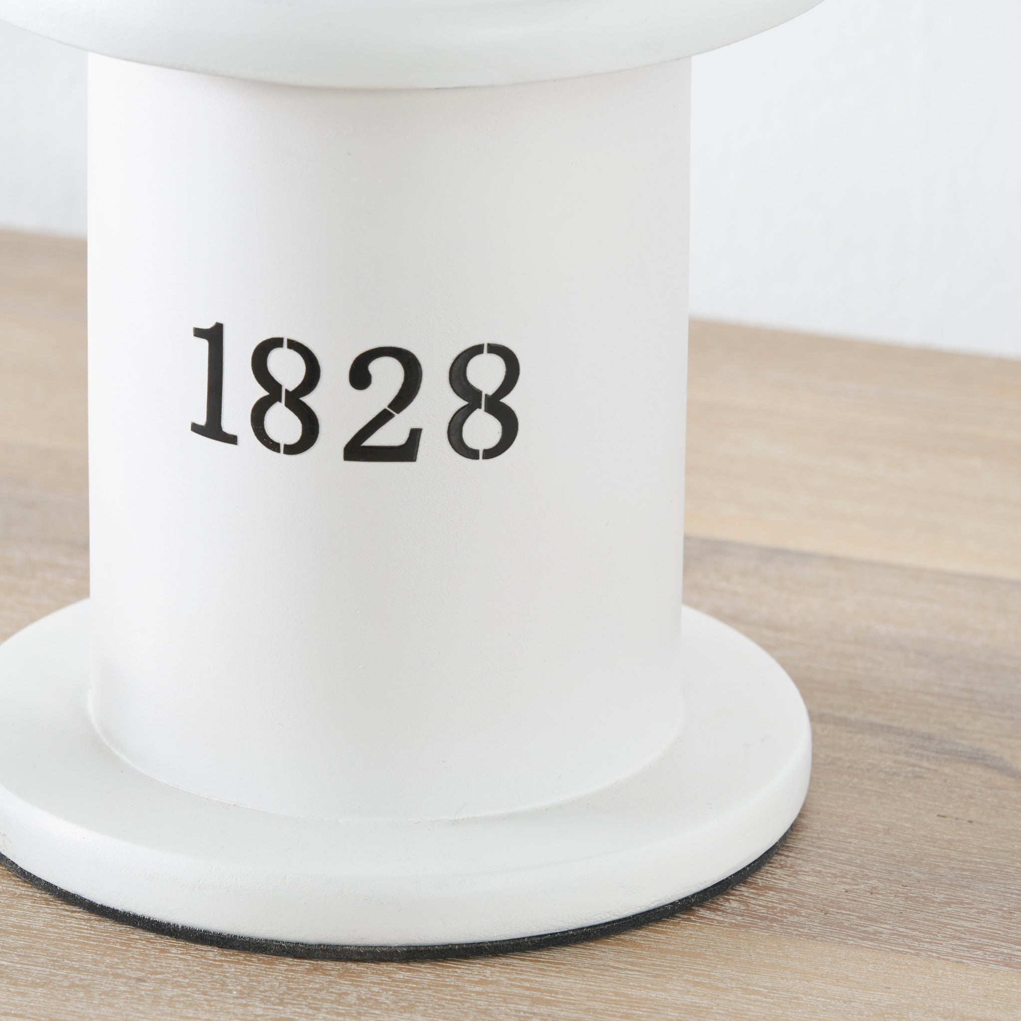 The Pacific Lifestyle Gibson Wood Lantern Table Lamp in white wood, featuring a cylindrical Victorian design and "1828" printed in black, rests on a wooden surface.