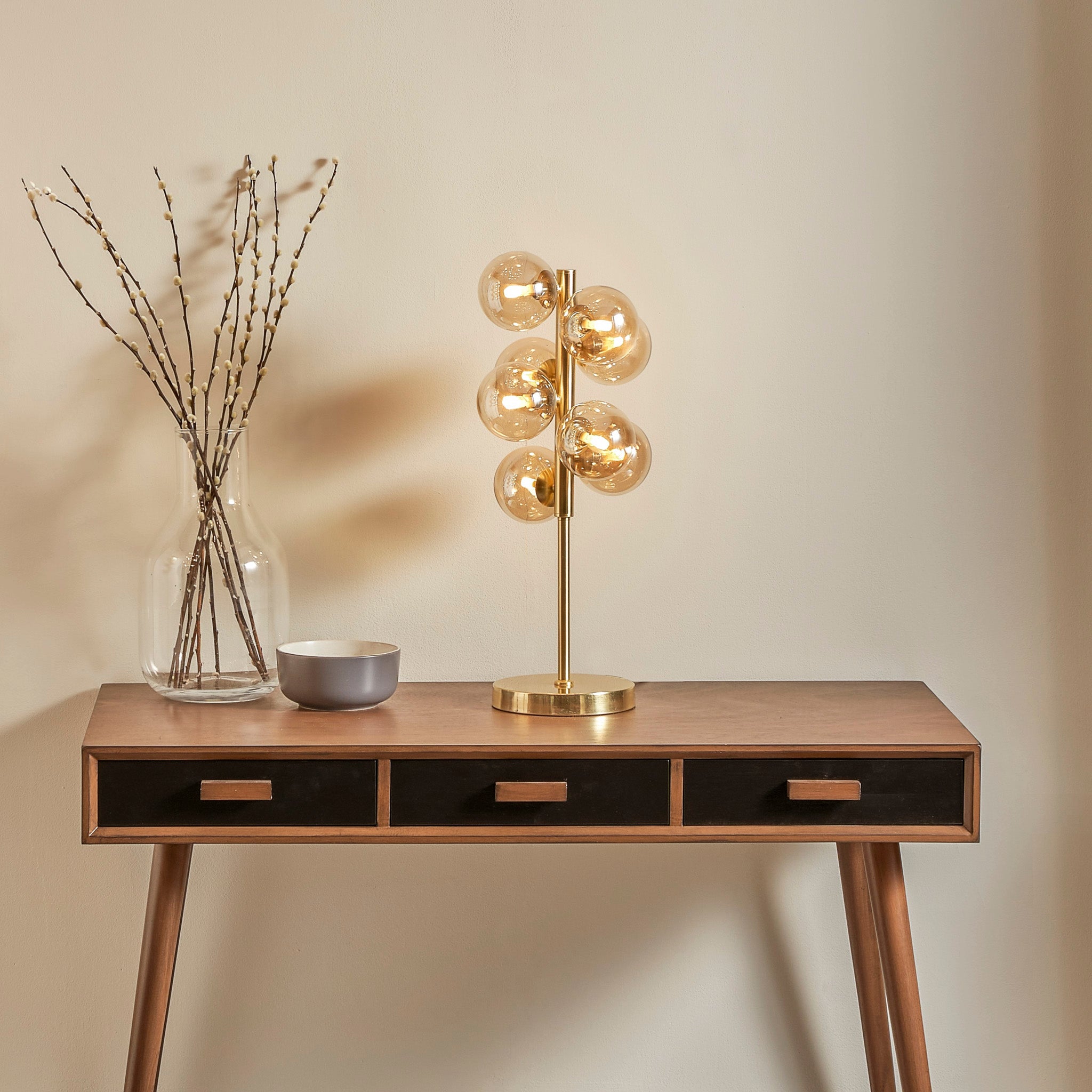 A modern table features the Pacific Lifestyle Vecchio Lustre Glass Orb and Gold Metal Table Lamp, sitting next to a clear vase with branches and a small gray bowl.