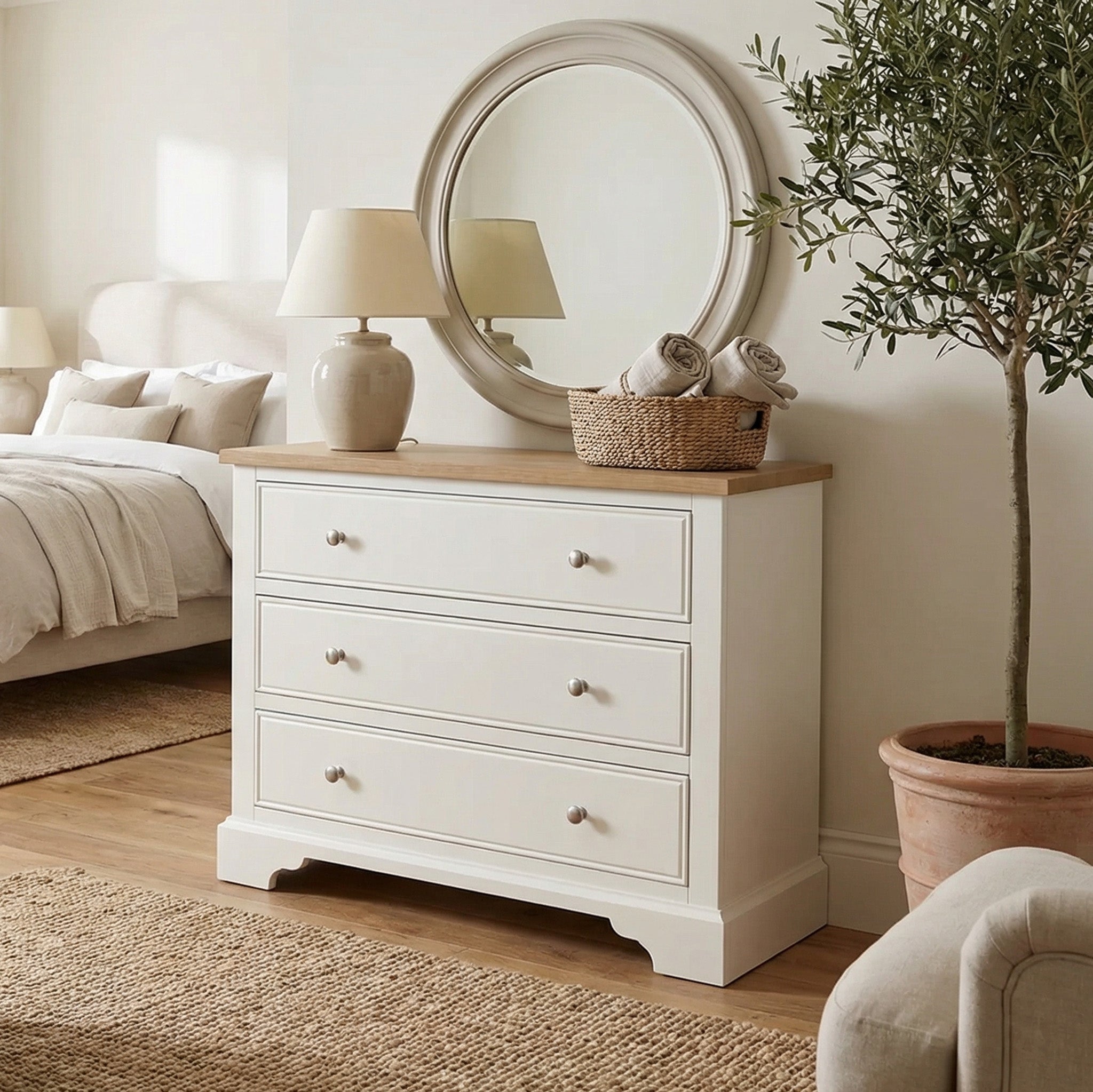 The Amberley 3 Chest of Drawers in Warm White, styled with a mirror, lamp, basket, and potted plant, provides compact storage for a cozy bedroom.